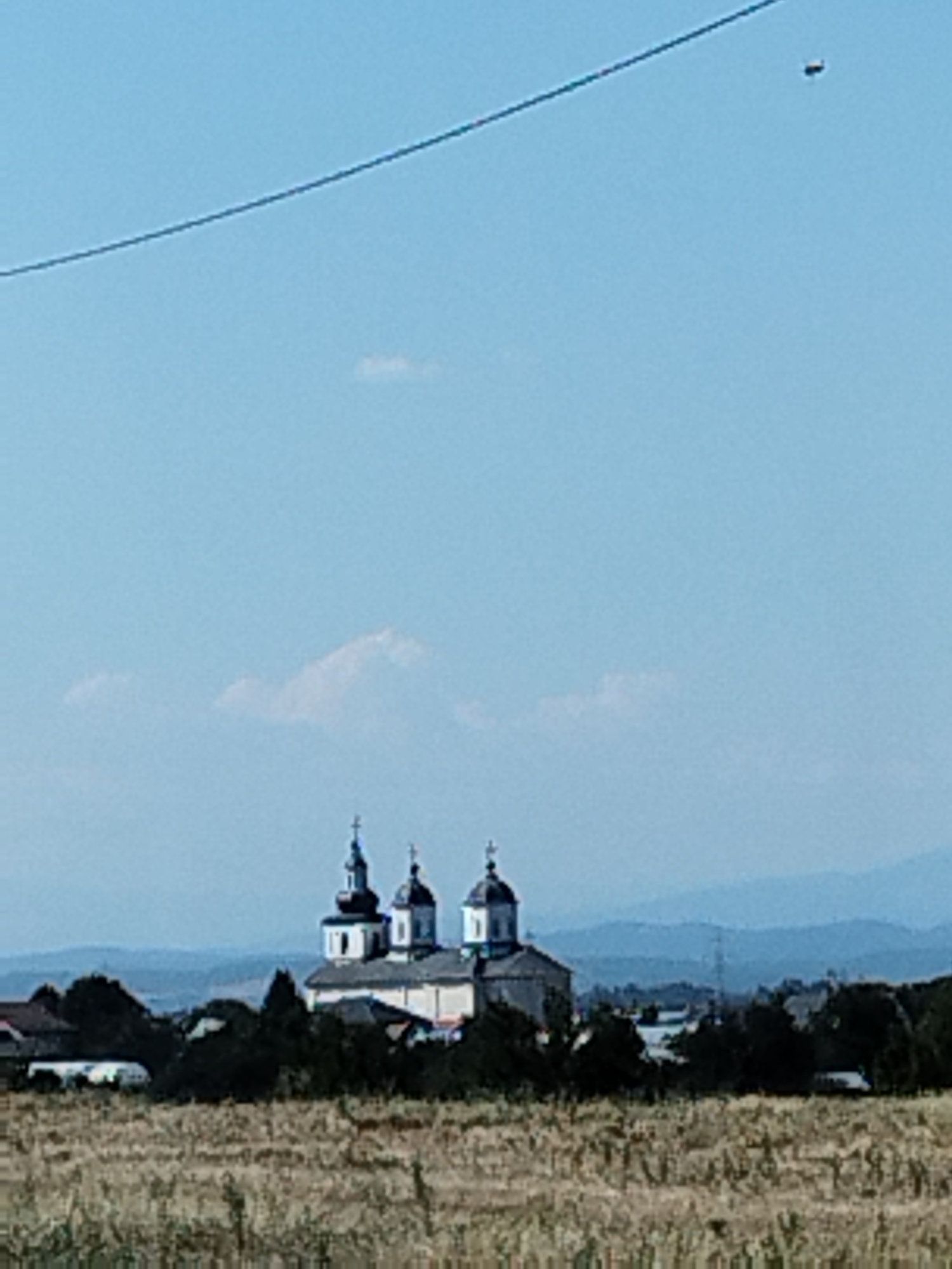 In Băleni-Români, Romania, a distant, low-resolution image captures a rural landscape under a bright, clear blue sky. In the middle ground, a church with multiple domes, characteristic of Orthodox architecture, stands prominently. It is a light-colored building with dark roofs and crosses atop its domes. Surrounding the church are scattered trees and buildings, suggesting a small village or hamlet. In the foreground, a dry, grassy field stretches across the lower portion of the image, indicating a warm, possibly late summer or autumn season. A thin, dark wire is visible diagonally across the upper left quadrant of the sky, and a small, dark, oblong object, possibly a drone or a distant bird, is present in the upper right quadrant of the sky. The background is a hazy landscape of rolling hills. The overall scene depicts a peaceful, rural setting.