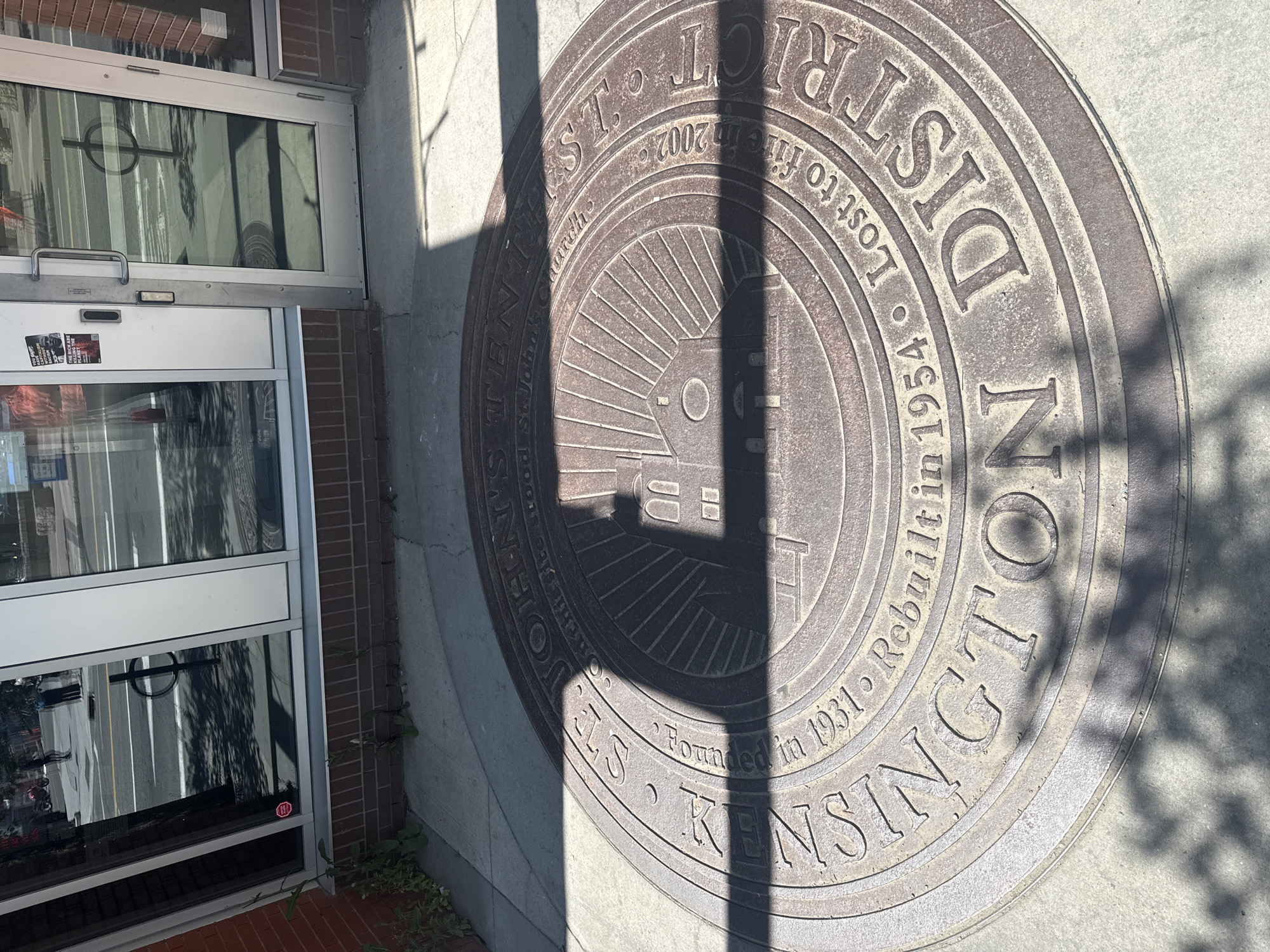 A circular bronze medallion embedded in a concrete sidewalk is the central focus of the image. The medallion displays text and an image. The text reads "St. John's Church Kensington District" and includes dates, "Founded in 1931", "Rebuilt in 1954", and "Lost to fire in 2002". The image on the medallion depicts a stylized church building with rays emanating from it. Sunlight casts a shadow of tree branches across the medallion and sidewalk. The sidewalk is adjacent to a building with large glass windows and brickwork at its base. In the reflection of the windows, a street scene is visible, with people and vehicles. The medallion is located in the Kensington District of Calgary, Alberta, Canada.