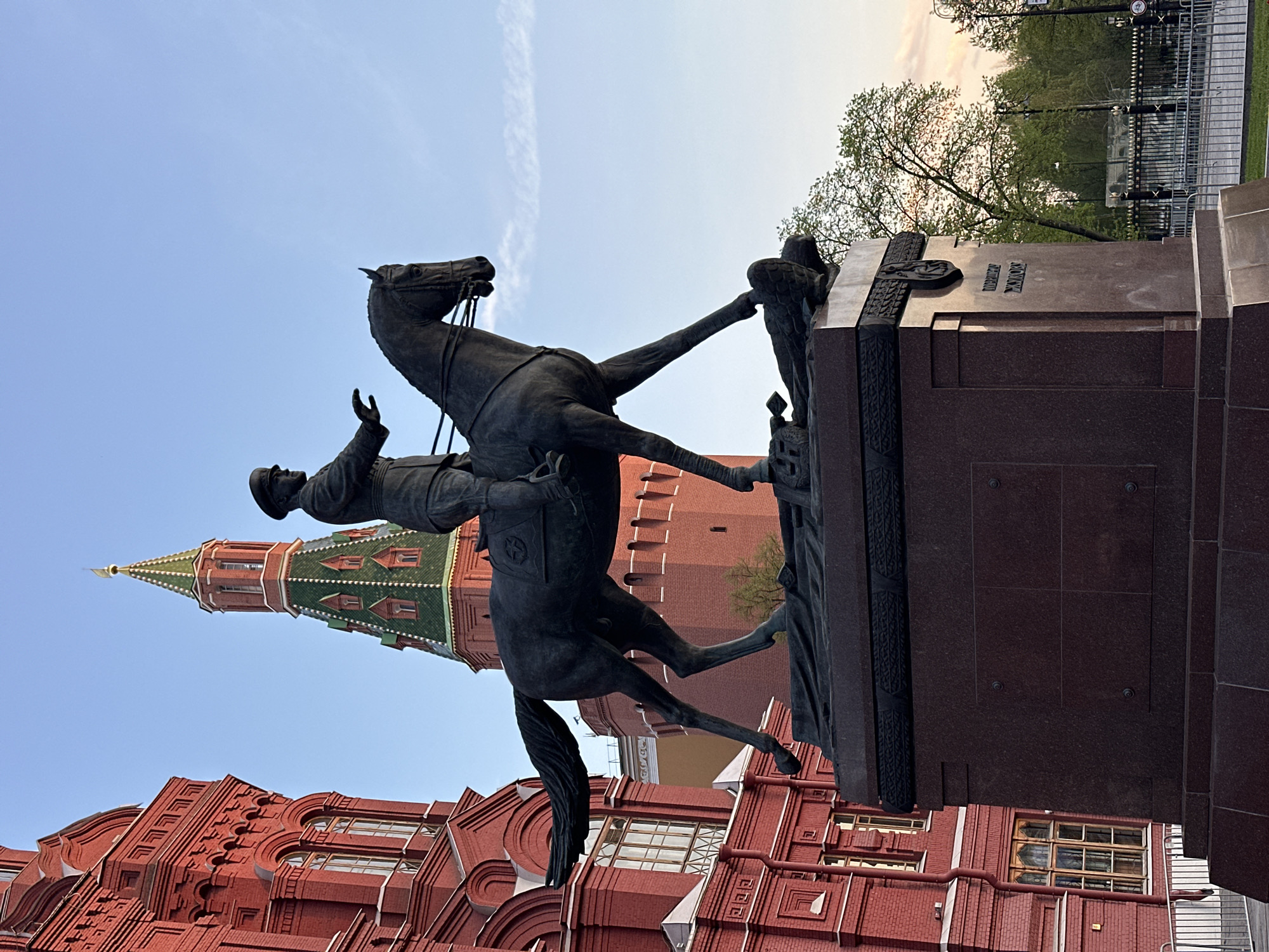 The image shows a bronze equestrian statue of Kuzma Minin and Prince Dmitry Pozharsky in Moscow, Russia, erected in front of St. Basil's Cathedral in Red Square. The statue depicts Pozharsky riding a rearing horse, with Minin raising his right arm. Behind the statue, the red brick architecture of the State Historical Museum and a tower of the Kremlin can be seen. The sky is blue with some clouds. Some trees are visible in the background.