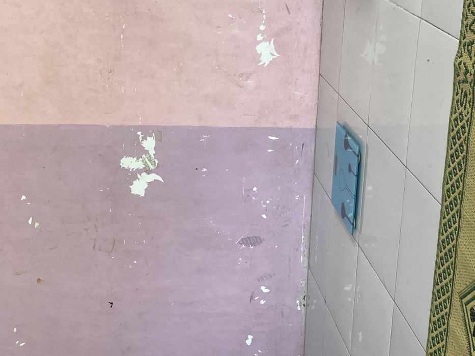 The image shows a bathroom scale, laying flat on a white tiled floor. The scale is blue and has black markings. The scale is situated in front of a wall that is painted in two shades of pink, separated by a vertical line. The wall has areas of peeling paint. The bottom of the image shows a portion of a green and brown rug. The location is a bathroom in Kecamatan Piyungan, Indonesia, at the coordinates provided.