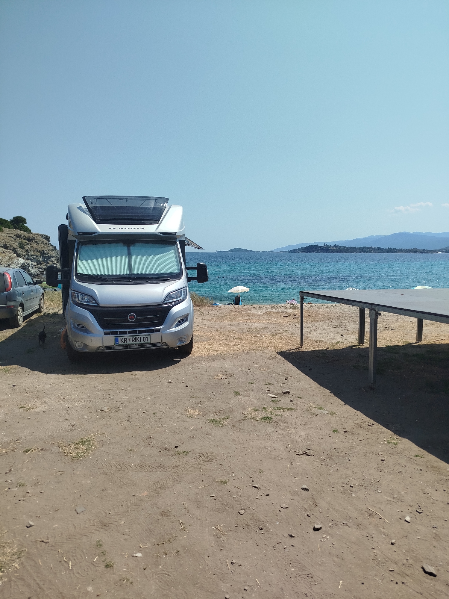 An Adria motorhome is parked on a sandy area near a beach. The motorhome is silver and has its front windshield covered by reflective shades. To the left of the motorhome is a silver passenger car, and a small black animal, possibly a cat, is walking near the front of the motorhome. In the background, the turquoise sea stretches to the horizon, with hills and mountains visible on the opposite shore. A white umbrella is set up on the beach, and a long, low table with metal legs is positioned in the foreground, casting a shadow. The overall scene depicts a serene, beachside camping location, likely in the Sithonia Municipal Unit of Greece, known for its coastal beauty.