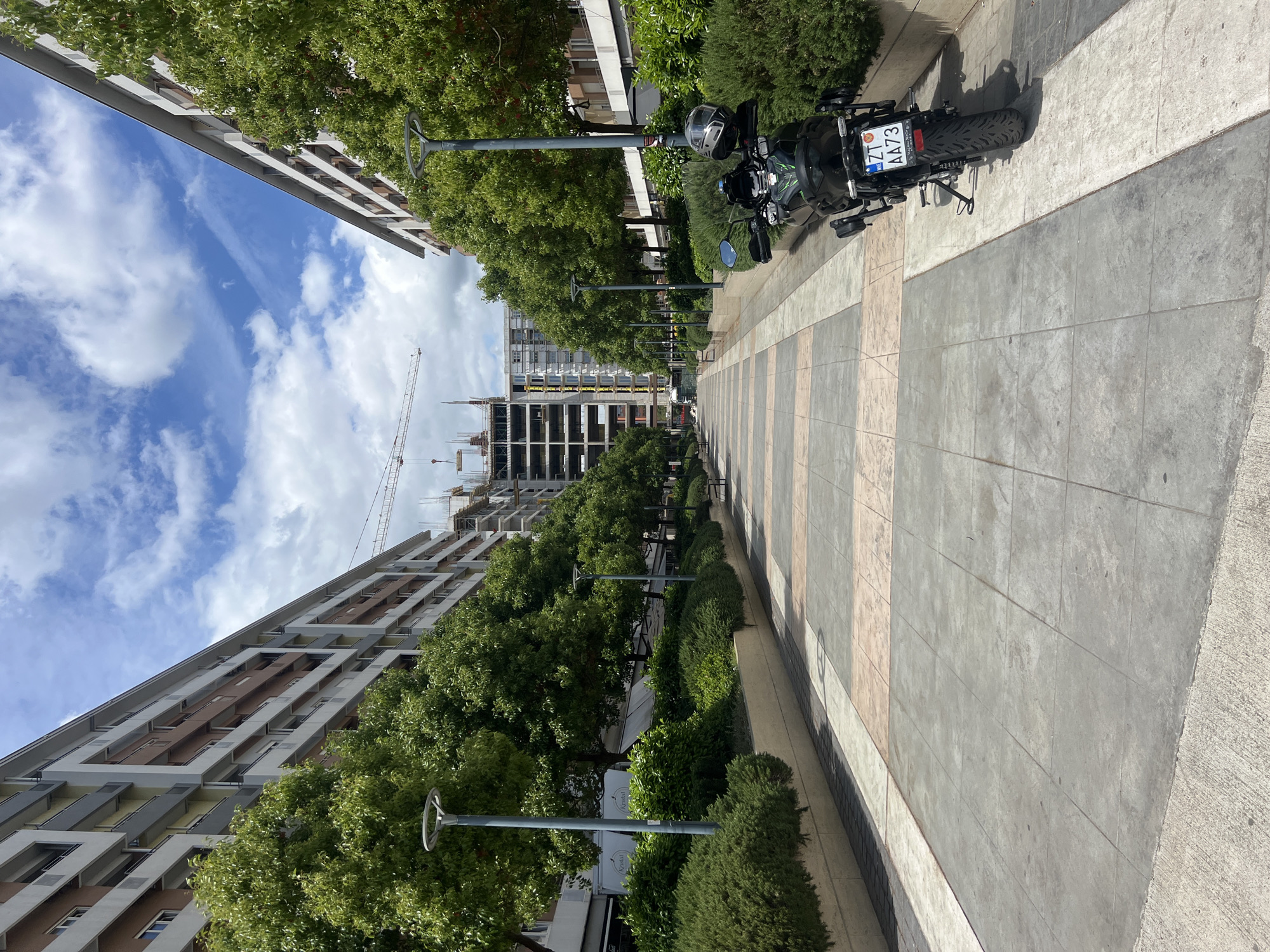 The image shows a straight pedestrian walkway in Podgorica, Montenegro, between two buildings, with trees and bushes lining the path. A motorcycle is parked on the right side of the walkway. In the distance, there is a building under construction with a crane. The sky is blue with some clouds. The walkway is paved with grey and beige tiles. Lampposts are visible along the walkway, interspersed among the trees. The motorcycle's license plate reads "ZT AA73".