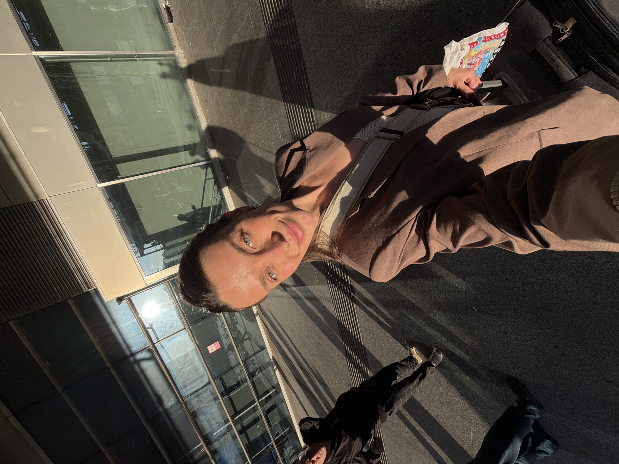 A woman in a brown suit with a cream-colored strap poses in a photograph taken at an extreme angle. She holds a magazine and stands near a modern glass-walled building, casting long shadows. The location is the city of Vnukovo, Russia. A partially visible figure is standing in the lower part of the picture.