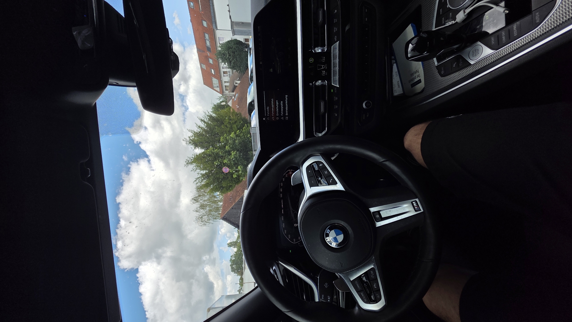 The image is taken from inside a car, looking out through the windshield. The car's dashboard, steering wheel, and gear shift are visible in the foreground. The steering wheel is black with silver accents and the BMW logo in the center. The dashboard features a digital display screen showing various vehicle information and menus. The gear shift is also visible, with the "START ENGINE STOP" button illuminated.
Outside the car, the scene depicts a partly cloudy sky with fluffy white clouds against a bright blue sky. Green trees with dense foliage are visible in the middle ground. Beyond the trees, buildings with red-tiled roofs and white walls are seen. The overall environment appears to be a suburban or semi-urban area on a sunny day. The image is a point-of-view shot, suggesting the photographer is the driver.