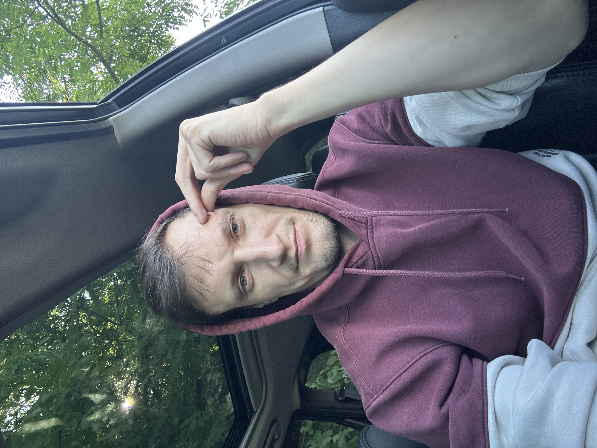 A man is sitting in a car, looking at the camera and touching his forehead with his right hand. He is wearing a maroon hooded sweatshirt over a cream-colored long-sleeved shirt. The car has a sunroof through which green trees and sunlight are visible. The location is Makiv, Ukraine.