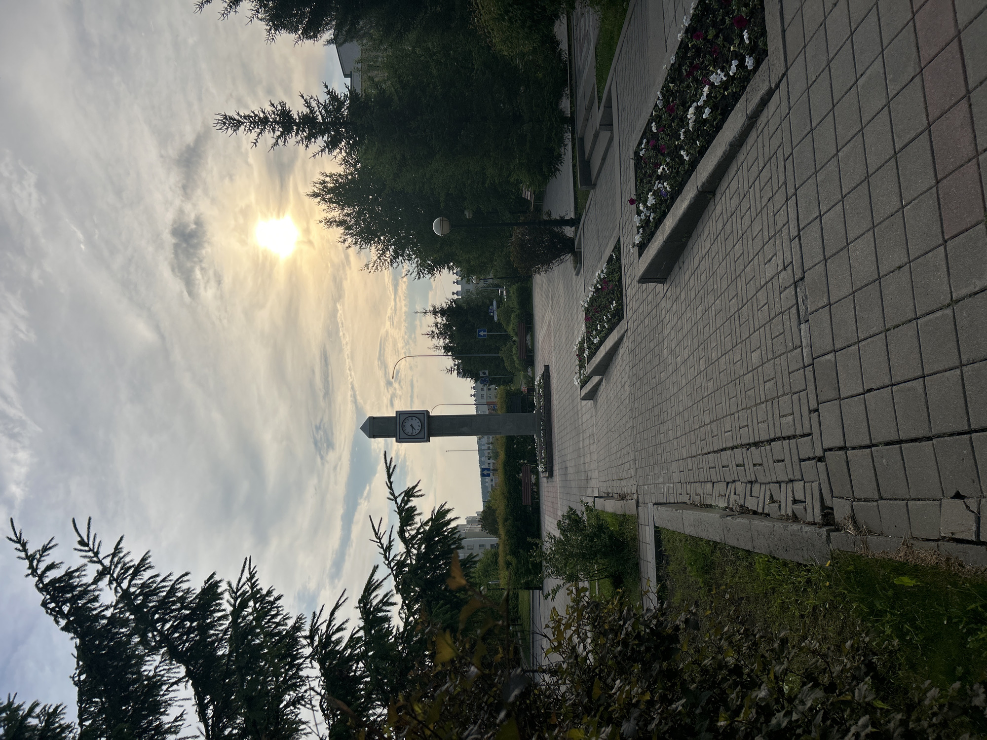 The image shows a paved walkway with a clock tower in the background. There are trees and flower beds lining the walkway. The sun is setting, casting a warm glow over the scene. The clock tower is made of stone and has a clock face on each side. The walkway is made of grey pavers and is in good condition. There are benches and streetlights along the walkway. The flower beds contain a variety of colorful flowers. The trees are evergreen and are tall and slender. The sky is cloudy with patches of blue. The overall scene is peaceful and serene.