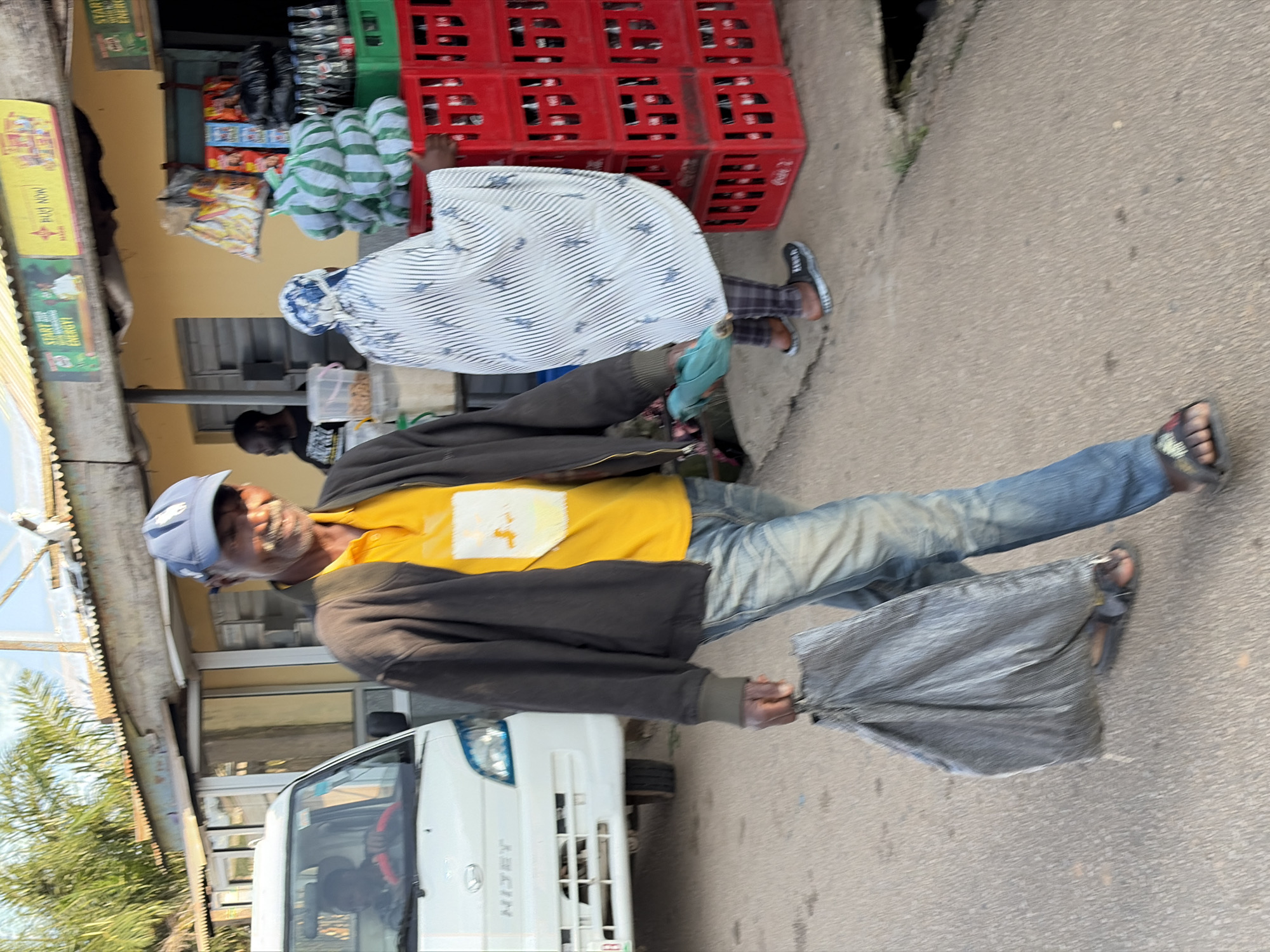 An elderly African man wearing a blue baseball cap, a yellow polo shirt, a dark jacket, and blue jeans walks down a paved street carrying a large, crumpled gray bag in his left hand. He is smiling and looking towards the camera. He is wearing sandals. Behind him, a white vehicle, possibly a Daihatsu Hijet, is parked. To his right, a woman is seen from behind, wearing a white and blue patterned headscarf and traditional clothing, standing next to stacks of red plastic crates filled with bottles, likely soft drinks. Behind the crates are shelves stocked with various products. The scene appears to be in front of a small shop or market. The overall lighting suggests it is daytime. The location is identified as Jos, Nigeria.
