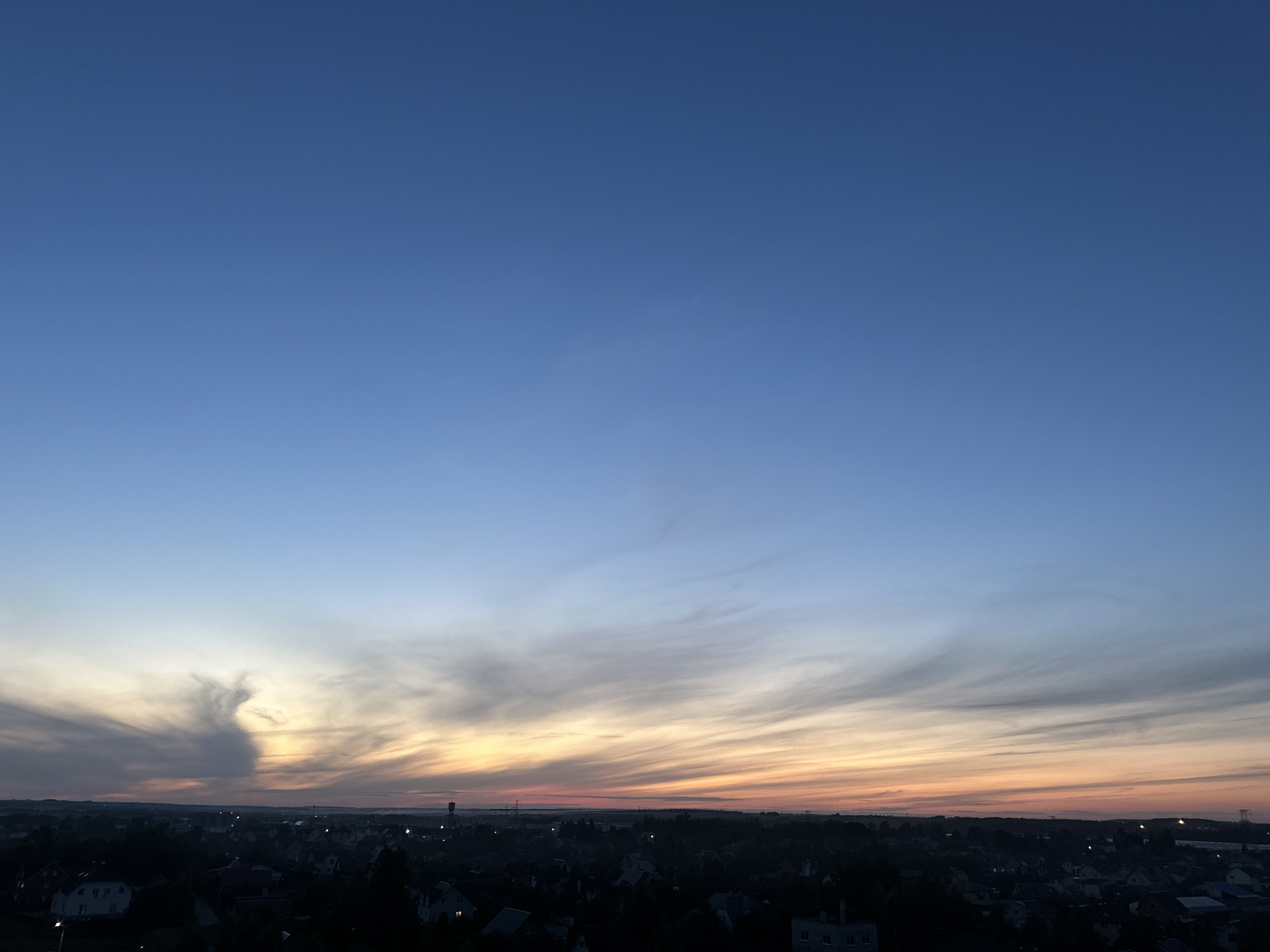 The image depicts a wide panoramic view of the sky above the settlement of Schomyślitski sel'ski Savec, Belarus, during twilight. The upper portion of the frame is dominated by a clear, deep blue sky that gradually transitions to lighter shades of blue and then to soft oranges and pinks near the horizon. Wispy, streaky clouds are scattered across the sky, particularly concentrated in the middle third of the image, catching the last rays of sunlight. These clouds create a dramatic, almost painterly effect.
Below the sky, a dark silhouette of a town or village is visible. The rooftops of numerous buildings are discernible, with some lights beginning to twinkle as darkness settles. The landscape appears to be relatively flat, with a few structures, possibly water towers or industrial elements, standing out faintly against the horizon. The overall impression is one of a peaceful evening setting, with the natural beauty of the sky contrasting with the developing urban landscape. There are no visible people or specific actions taking place.