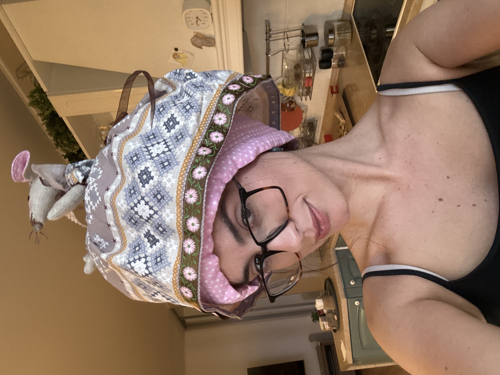 A woman wearing a patterned bonnet and glasses is oriented towards the viewer in an indoor setting, possibly a kitchen. The woman is wearing a black tank top with a white and black strap. The background includes kitchen cabinets, a stove top, and various kitchen utensils and appliances. The location appears to be a residential space, but the exact city and area are not discernible from the image.
