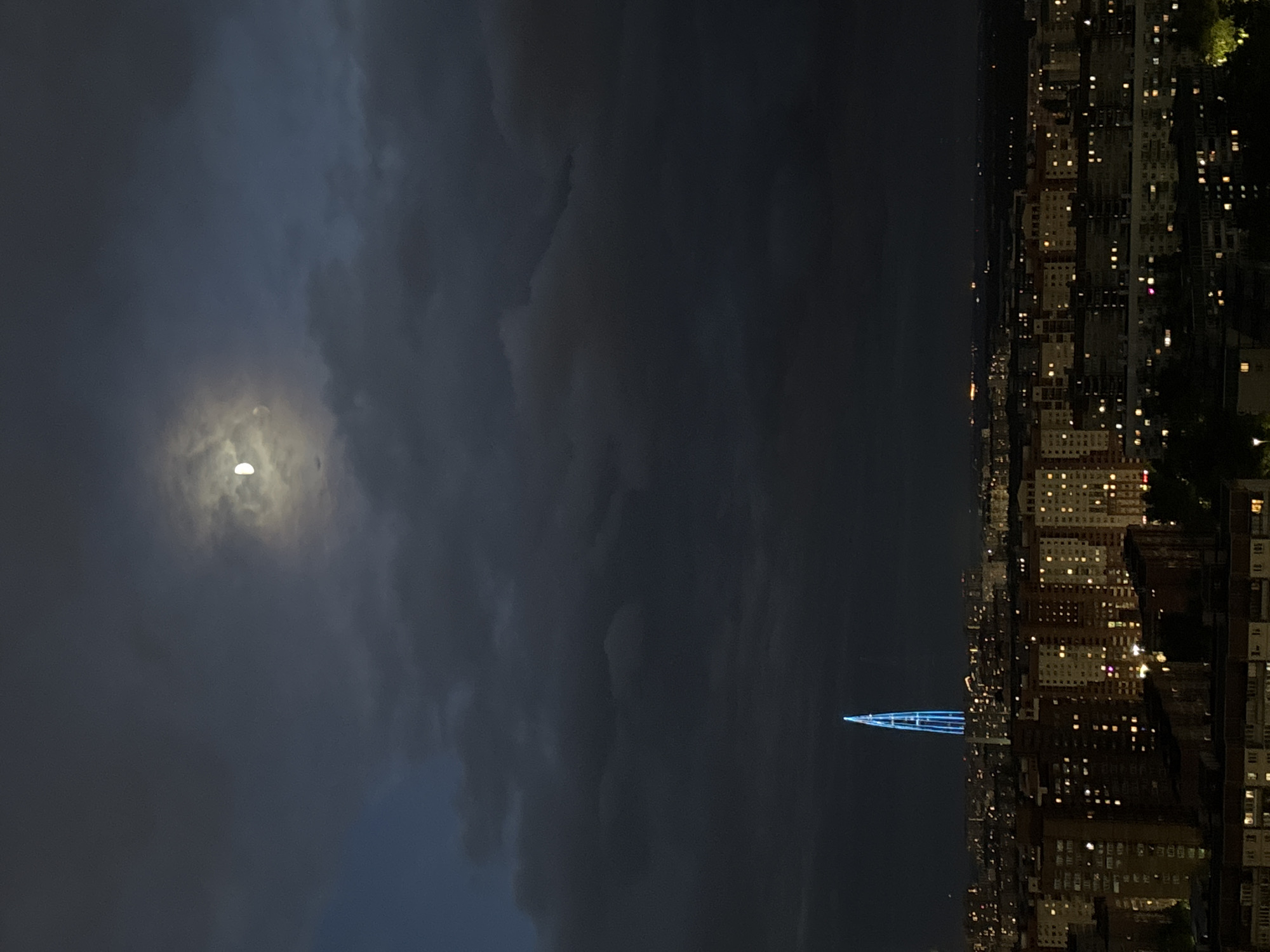The image shows an evening cityscape in Saint Petersburg, Russia. On the left, a partially obscured moon shines through a cloudy, dark sky. On the right, a densely populated urban area is visible. Buildings with illuminated windows line the horizon. In the center, a structure with a blue-lit, pointed peak stands out among the buildings.