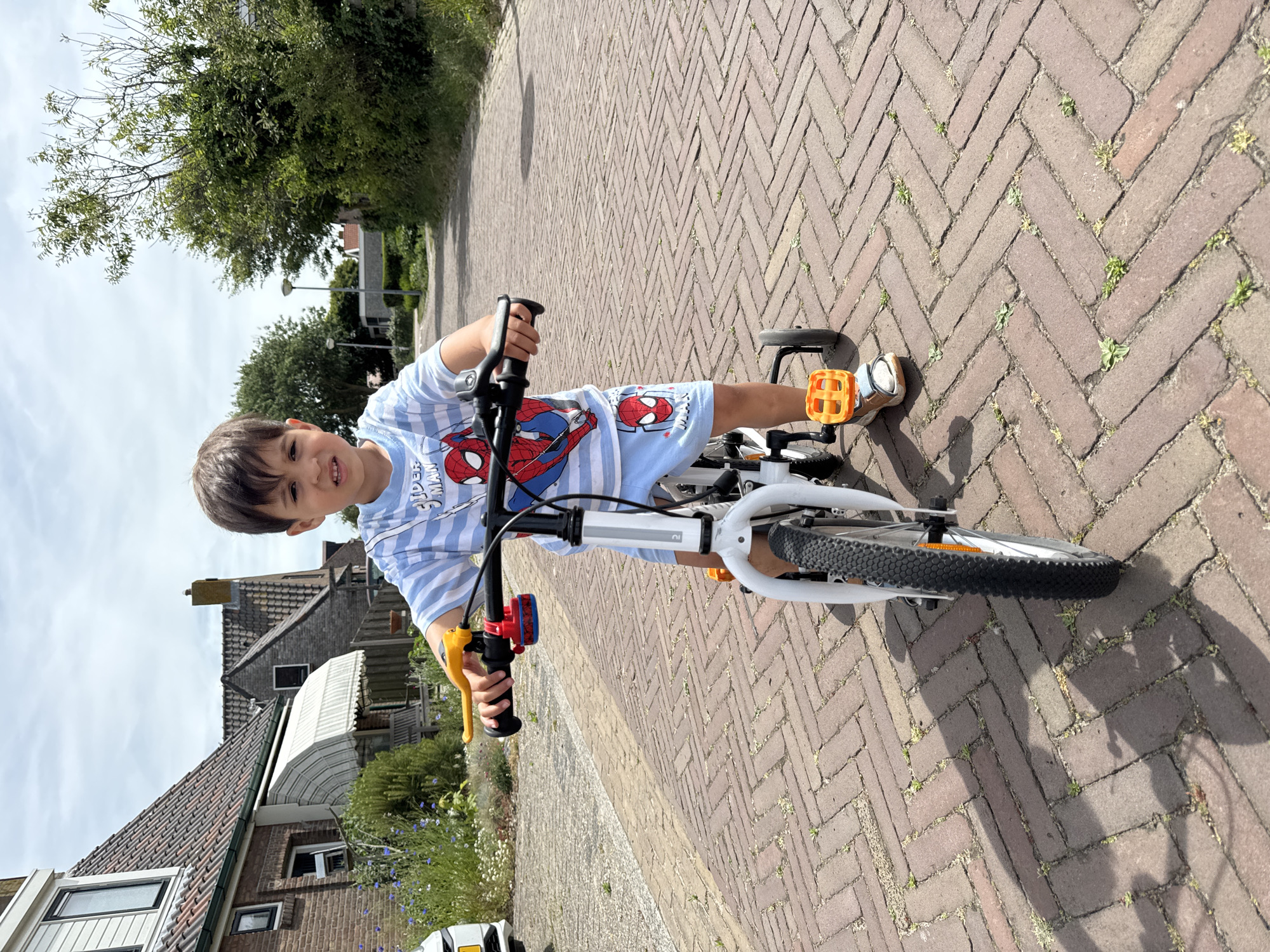 A young boy rides a white bicycle with orange pedals on a brick-paved street. The boy is wearing a blue and white striped shirt with a Spiderman graphic and matching shorts. He looks at the camera, holding the handlebars. The location is Petten, Netherlands. Houses are visible in the background, along with trees and other vegetation.