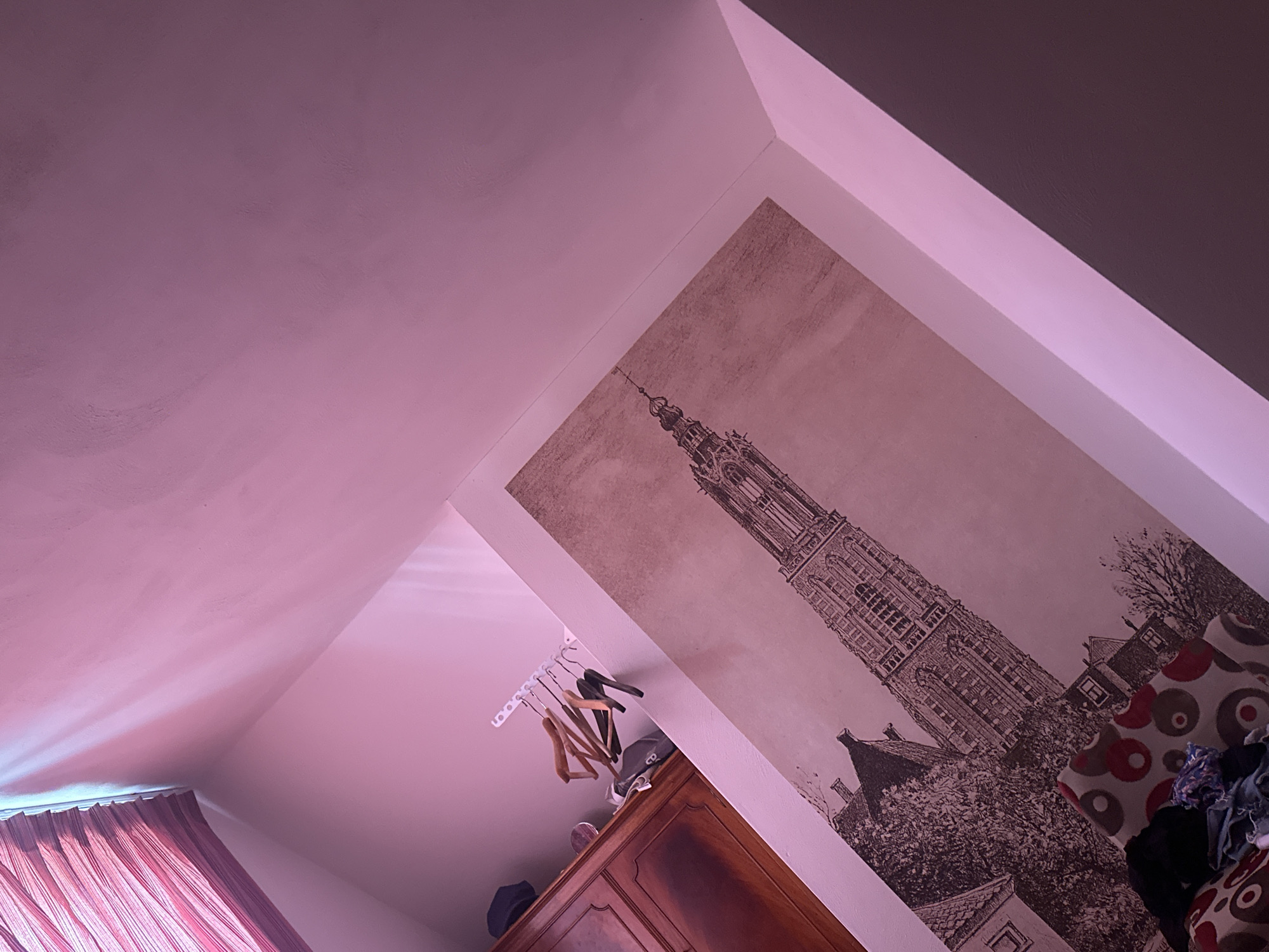 The image shows the interior of a room with an angled ceiling. The walls are painted a light shade of pink. On one wall, there's a large print of a building, reminiscent of the Onze Lieve Vrouwetoren, a church tower located in Amersfoort, Netherlands. A wooden cabinet sits beneath the print, with clothes hangers mounted on the wall above. A partially visible red curtain is on the lower-left side of the image. There are pillows on the floor next to the building.