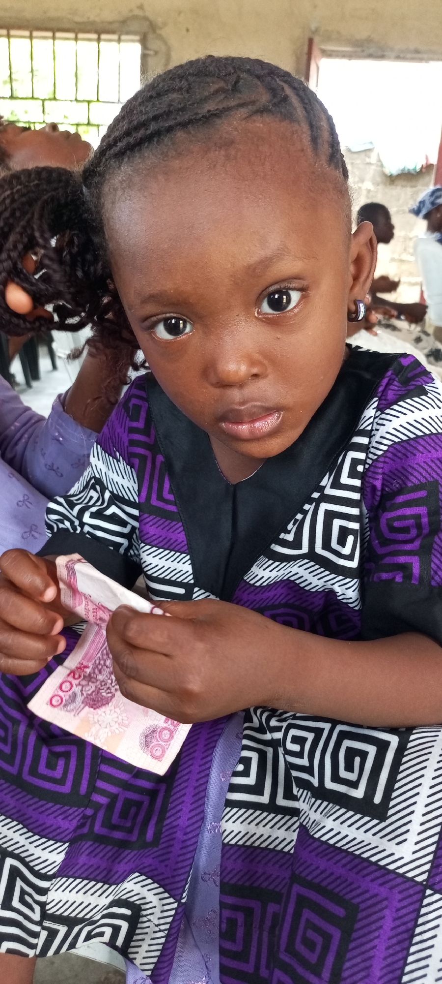 A young girl is indoors in Unknown, Unknown, holding two 100 yuan Chinese banknotes. She is sitting down and looking directly at the camera. She is wearing a purple, black, and white patterned shirt with a black collar and a silver hoop earring in her left ear. Her hair is styled in neat braids. To her left are the braids of another person, and in the background are other individuals. The environment appears to be a simple indoor setting.
