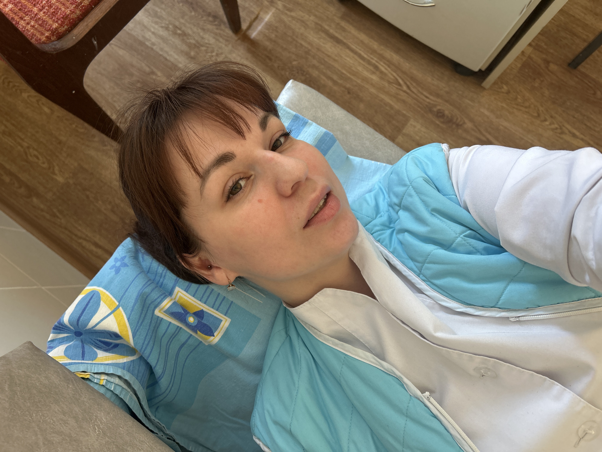 A female individual with medium brown hair and light-colored eyes is depicted in a reclined position, looking directly at the camera. She is wearing a white collared garment under a light blue quilted vest. A piercing is visible on her upper left ear, along with a thin, dangling earring. She is situated on a light-colored cushioned surface covered by a blue fabric sheet patterned with abstract blue and yellow motifs. The immediate background shows wood-look flooring, a partial view of a dark brown wooden chair with a red upholstered seat, and a light-colored, possibly electronic, object on the floor. The environment suggests an indoor setting, potentially a private room or facility. The location in the city of Фоки, Russia, cannot be confirmed by visual analysis of the image.