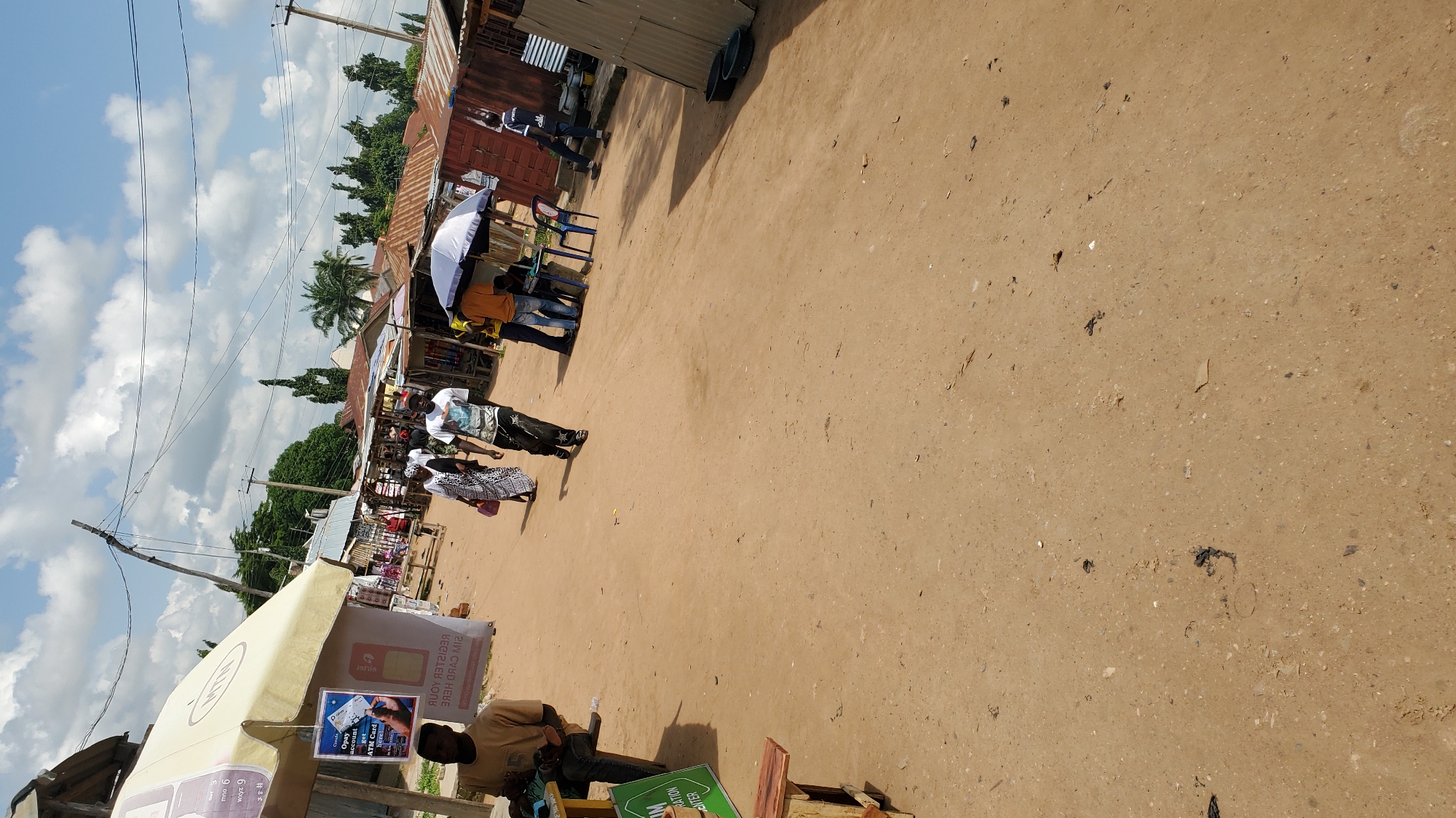 In Jalingo, Nigeria, under a partly cloudy sky, a dirt road bustles with activity. A man in a tan shirt and dark pants sits on a wooden bench to the left, seemingly observing the scene. Further down the road, a woman in a patterned dress walks, followed by a man in a white t-shirt and dark pants. They approach a cluster of market stalls with corrugated metal roofs. At one stall, a blue and white umbrella shades a table where three men are gathered, two standing and one seated. To the right, another man in a dark shirt walks away from the stalls. The ground is a mixture of sand and scattered debris. Overhead, power lines crisscross the sky, supported by wooden poles.