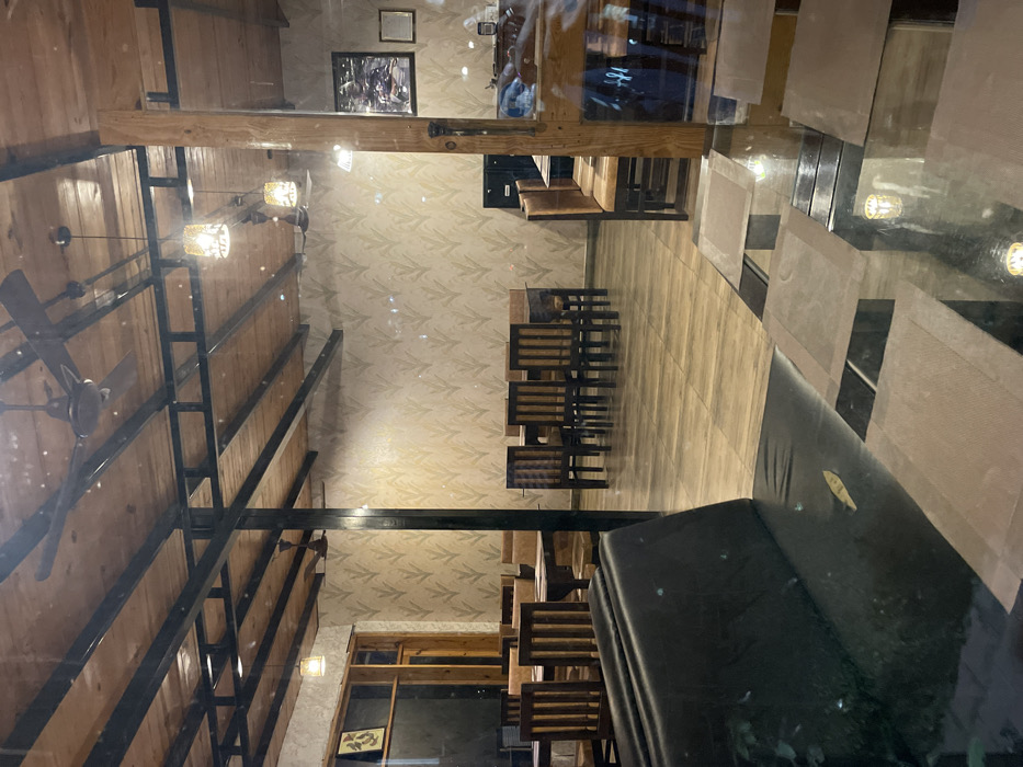 This image presents the interior of what appears to be a restaurant or a dining facility, consistent with a setting in Bhimtal, India. The architecture combines rustic and contemporary elements.
The prominent feature is a high, sloped ceiling crafted from light-toned wooden planks, reinforced by dark, exposed metal beams. Multiple dark ceiling fans are mounted on this wooden ceiling, alongside several ornate pendant lights with textured glass shades, casting a warm, inviting glow throughout the space.
One wall is covered with a subtle, light-colored wallpaper featuring a repeating leaf pattern, adding a natural touch to the decor. This wall also displays framed artwork, including a larger landscape picture and a smaller, more abstract piece. The flooring transitions between light, wood-effect tiles or planks and a darker, reflective surface in the foreground.
The furniture includes several sets of dining tables accompanied by wooden slatted chairs, suggesting a capacity for multiple diners. Further into the room, a wooden counter or bar area is visible, complemented by booth-style seating. In the immediate foreground, a plush, dark upholstered sofa or bench offers lounge-style seating.
The restaurant is currently unoccupied, with no people visible, implying the photograph was taken during off-peak hours or before service. The predominant use of artificial lighting suggests it is either evening or night. The overall impression is one of a well-maintained, tranquil, and tastefully decorated establishment, ready to welcome guests. There is no visible text in the image.