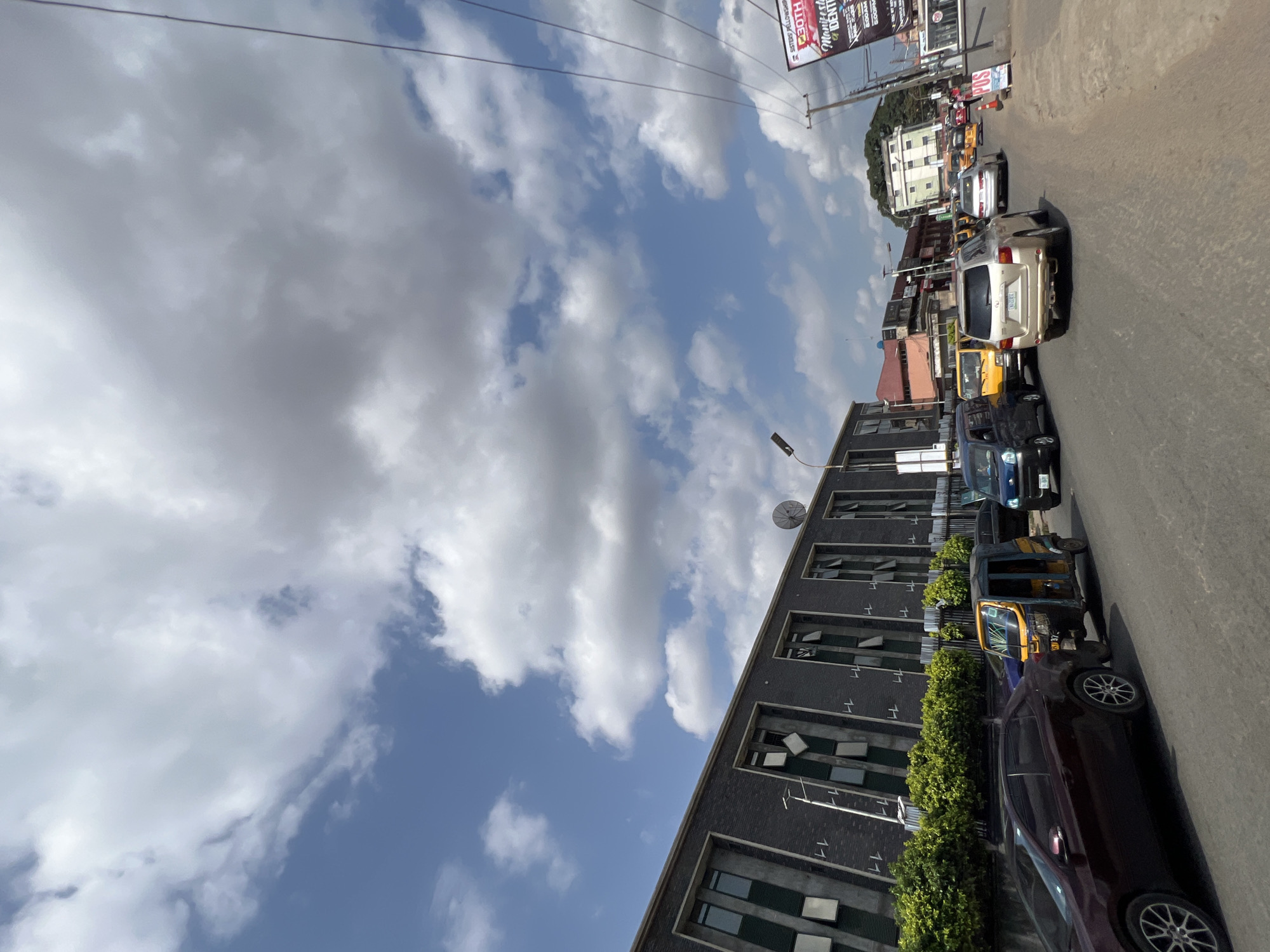 This image captures a vibrant urban street scene in Onitsha, Nigeria, under a bright, partly cloudy sky. The photograph is tilted clockwise, presenting the ground and buildings at an angle.
In the upper left, a expanse of clear blue sky is visible, interspersed with large, fluffy white cumulus clouds, indicating a sunny day. Utility lines stretch across the upper portion of the frame.
On the left side, a multi-story modern building with a dark, possibly brick or panelled, facade runs parallel to the street. It features numerous rectangular windows, some of which appear open or are designed with a louvered effect. A white satellite dish is mounted on the building's roof. A line of green, well-maintained bushes forms a low hedge along the front of the building, separating it from the road.
The right side of the image depicts an asphalt road with a moderate flow of vehicles, suggesting either traffic or roadside parking. In the immediate foreground, a dark red or maroon sedan is partially visible. Behind it, a yellow and black Keke Napep (motorized tricycle, a common form of public transport in Nigeria) is stopped or parked. Further along the road, a blue car, a gold-colored SUV (likely a Lexus), and another yellow Keke Napep are visible. A white van or SUV and other vehicles can be seen in the distance, indicating a busy thoroughfare.
Several commercial billboards are present further down the road on the right. Partial text visible on one large billboard includes "SISTERS FELLOWSHIP", "30TH", "MANIFEST THE IDENTI", and "ECOBANK". Another smaller sign reads "P.I.S". The overall impression is a bustling commercial area, characteristic of a city environment.