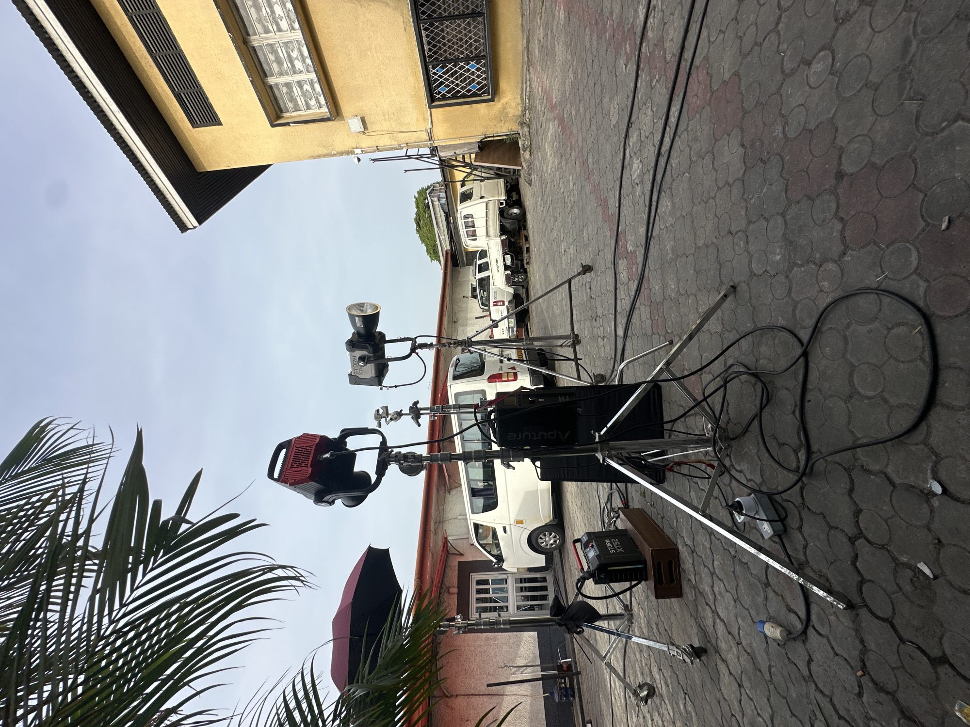 The image captures an outdoor film or photography set in Lagos, Nigeria. The foreground features lighting equipment on stands, including a red LED panel light, a reflector, and an umbrella. There are power supplies and cables strewn across the ground, which is paved with a patterned gray brick. In the background, there are several white vans parked in a narrow alley between two buildings. One building is painted yellow, with security bars on the windows. The other building is painted red. A palm tree branch is visible on the left side of the frame. The sky is overcast.