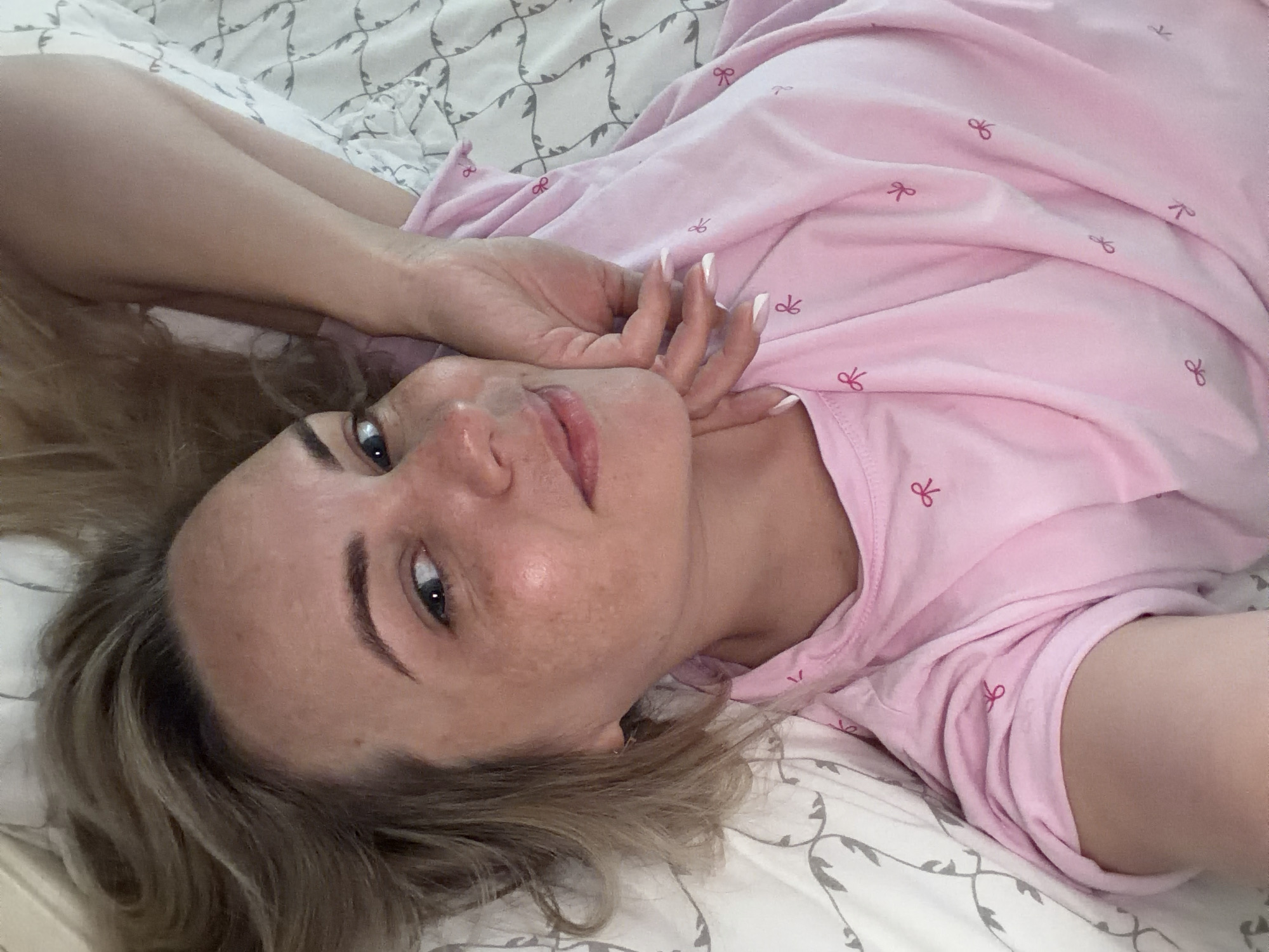 A medium-close up shot depicts a woman lying on her back on a bed, looking directly at the viewer with a slight tilt of her head. Her expression is relaxed and neutral, with a hint of a gentle smile. She has blonde or light brown hair fanned out on the white and grey patterned bedding.
The woman is wearing a light pink top adorned with small, darker pink bow patterns. Her left hand, featuring long nails with white tips (a French manicure style), is gently placed near her jawline and neck. Her visible eye is blue or grey, and her eyebrows appear well-defined. Freckles are visible on her cheek and nose. Her lips are slightly parted.
The setting is indoors, likely a bedroom, characterized by the light-colored bed linens with a subtle, swirling grey pattern. The lighting suggests an indoor environment, without strong shadows or bright natural light. There is no visible text in the image. The scene is set in Votkinsk, Russia.