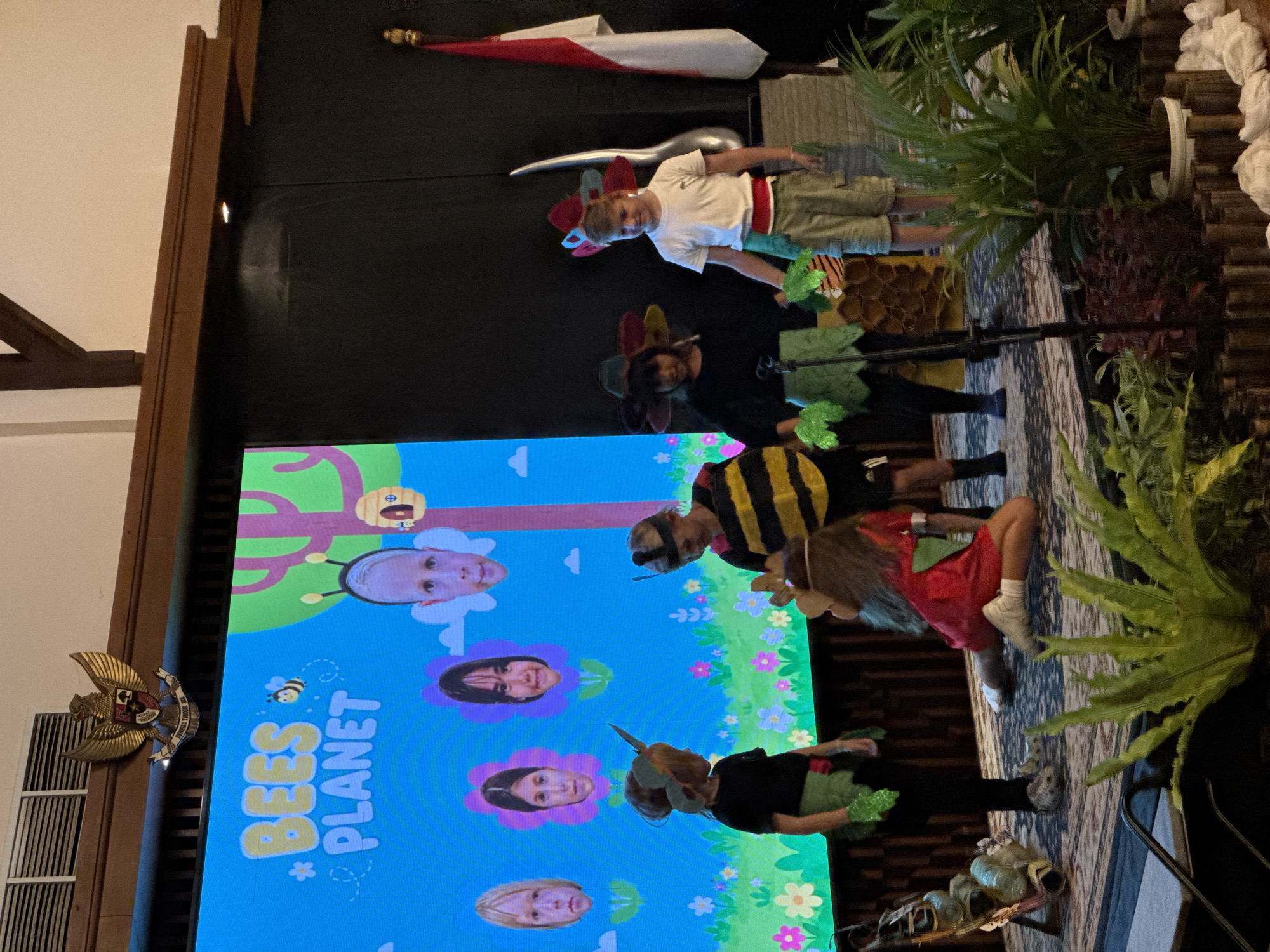 An indoor event takes place on a stage in Kuta Selatan, Indonesia, with a large LED screen displaying a colorful background with cartoon bees and the text "BEES PLANET" and flower graphics. In front of the screen, several children are on stage, some wearing costumes. One child in the foreground, on the right, wears a bee costume with black and yellow stripes and a bee headband. Another child, in the center, is kneeling and wearing a red dress and green leafy accessories. To the left, another child stands, dressed in green, with a flower-like headdress. Behind them, several other children are visible, some with flower-themed costumes. The stage is decorated with plants and what appears to be a wooden honeycomb structure. A national flag of Indonesia, a white and red bicolor, is displayed to the right of the stage. Above the screen, a crest is mounted on the wall. The overall scene suggests a children's performance or presentation related to bees and nature.