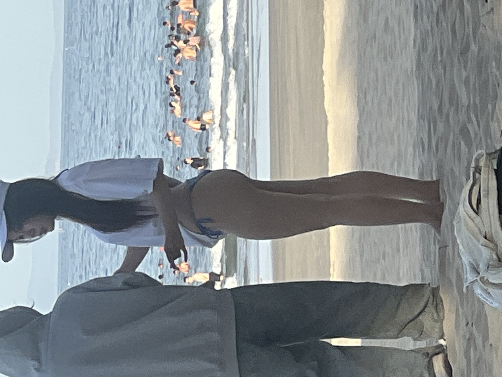 The image depicts a group of people on a beach, with two individuals prominently in the foreground, facing away from the camera. The person on the left wears a grey hooded sweatshirt and dark pants, while the person on the right wears a white t-shirt and a blue bikini bottom, with a white baseball cap. They appear to be standing and interacting. In the background, the ocean is visible with numerous people swimming and playing in the water. The beach itself is sandy. The lighting suggests it is daytime, possibly late afternoon given the soft light. The scene is generic and does not contain any specific landmarks or indicators that would identify it as being in the city of Đà Nẵng, Vietnam.