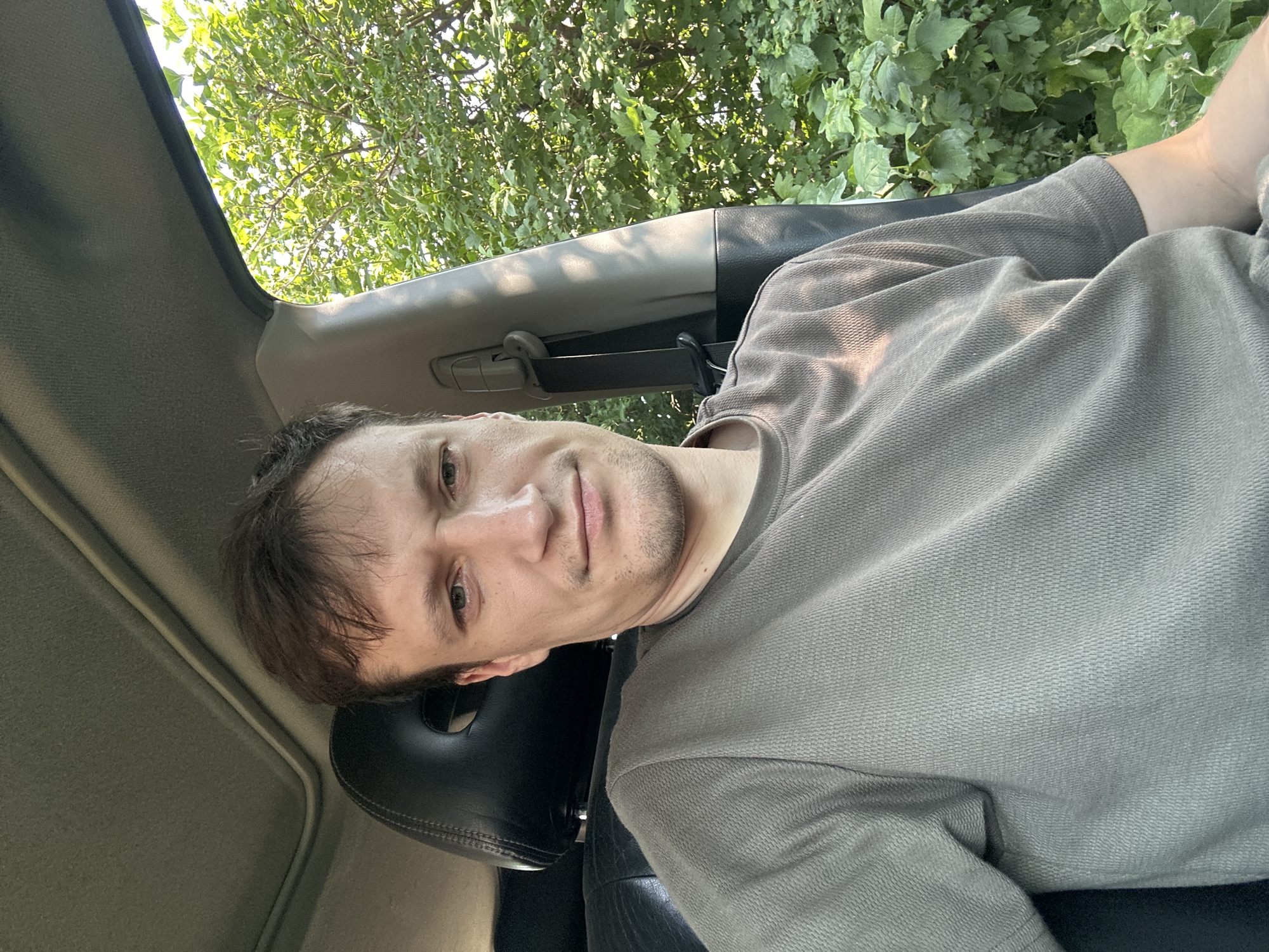 A man with short brown hair and light skin is sitting in the driver's seat of a car, looking at the camera. He is wearing a grey t-shirt. The car's interior is visible, including a black leather headrest and a sunroof. Outside the car, green trees and foliage are visible. The location is Shatava, Ukraine.