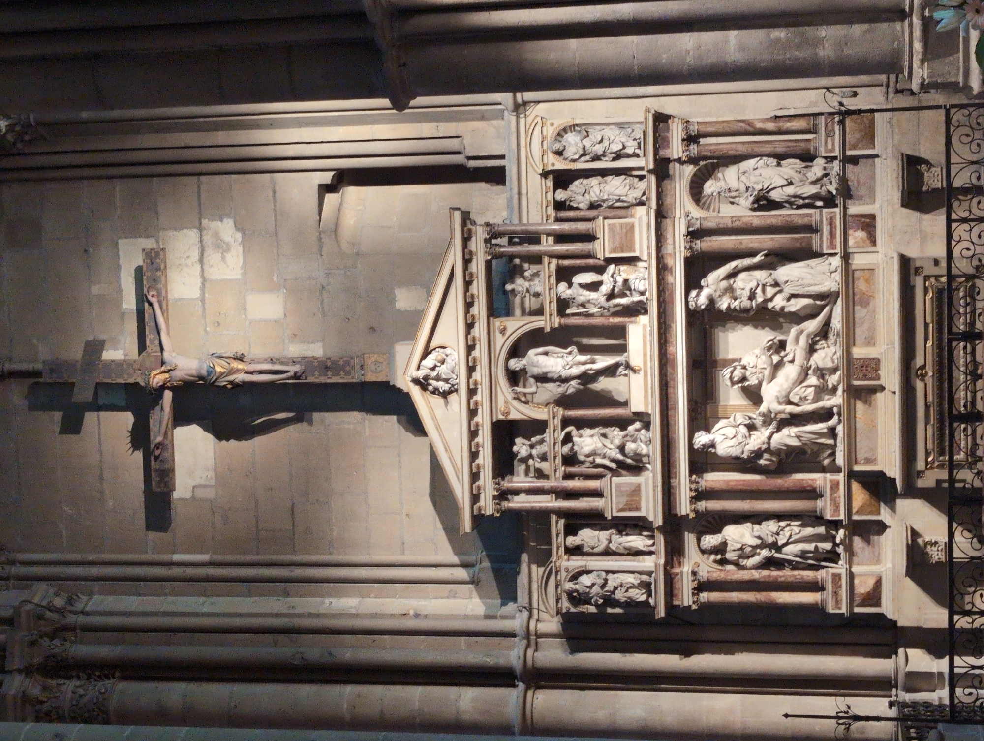 The image displays a detailed sculpture of a crucifixion scene within a church in Urvillers, France. The central element is a large wooden cross with a sculpture of Jesus Christ affixed to it. To the right of the cross, a shadow is cast onto the stone wall of the church. Below the crucifixion, a complex multi-tiered marble monument is present, adorned with numerous sculpted figures. These figures depict various religious scenes and individuals, including what appears to be a pietà on the lower tier. The monument features architectural elements like columns and pediments. The overall setting is that of a dimly lit church interior, with stone columns visible on the left and right edges of the frame, and elaborate stonework forming the backdrop. A black metal railing is visible at the bottom of the image, suggesting a roped-off area in front of the monument.