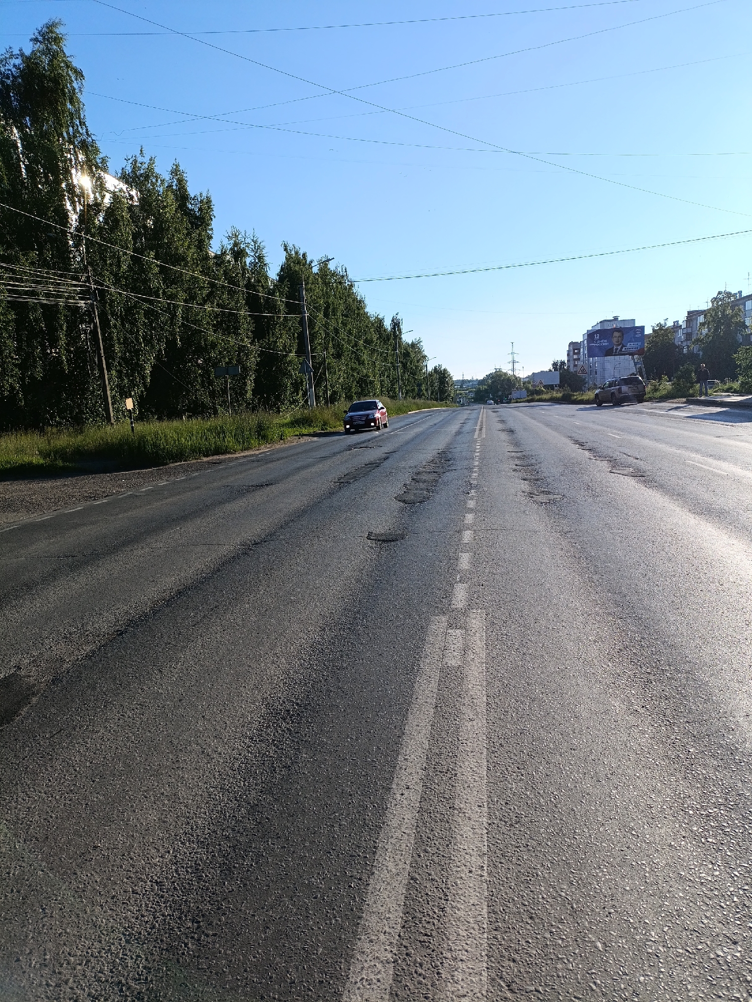 The image depicts a wide, asphalt road in Chaikovsky, Russia, under a clear blue sky. The road is lined with trees on the left and buildings and a billboard on the right. The asphalt surface is uneven, with several potholes visible. A red car is driving on the road in the left lane, while a darker colored SUV is parked on the right side of the road. A person is standing near the SUV. Power lines are visible strung across the sky. The sun is shining brightly, casting shadows and highlighting the textures of the road surface.