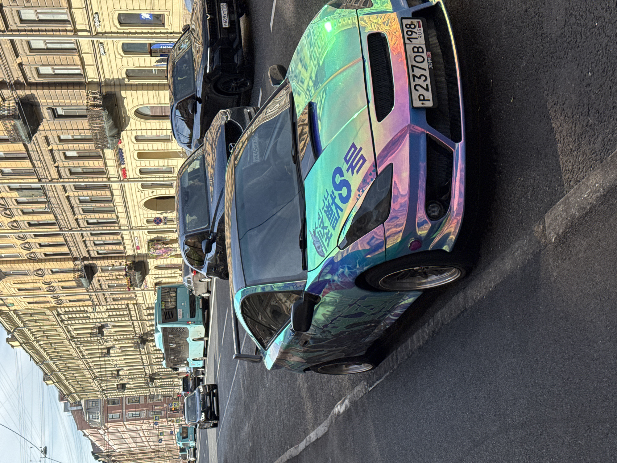 This image depicts a street scene in Saint Petersburg, Russia, likely on a sunny day. In the foreground, a highly customized car with a holographic, iridescent wrap is parked on the right side of a multi-lane road. The car features a large rear spoiler and a modified front bumper. Its license plate reads "P237OB 198 RUS".
In the background, several vehicles, including a modern black SUV and two light blue buses, are driving on the road. The buildings lining the street are multi-story, with ornate architectural details characteristic of Saint Petersburg. The buildings have numerous windows, some with balconies, and appear to be shops or residences. Overhead, power lines and streetlights are visible. The overall atmosphere suggests a typical urban environment in the city.