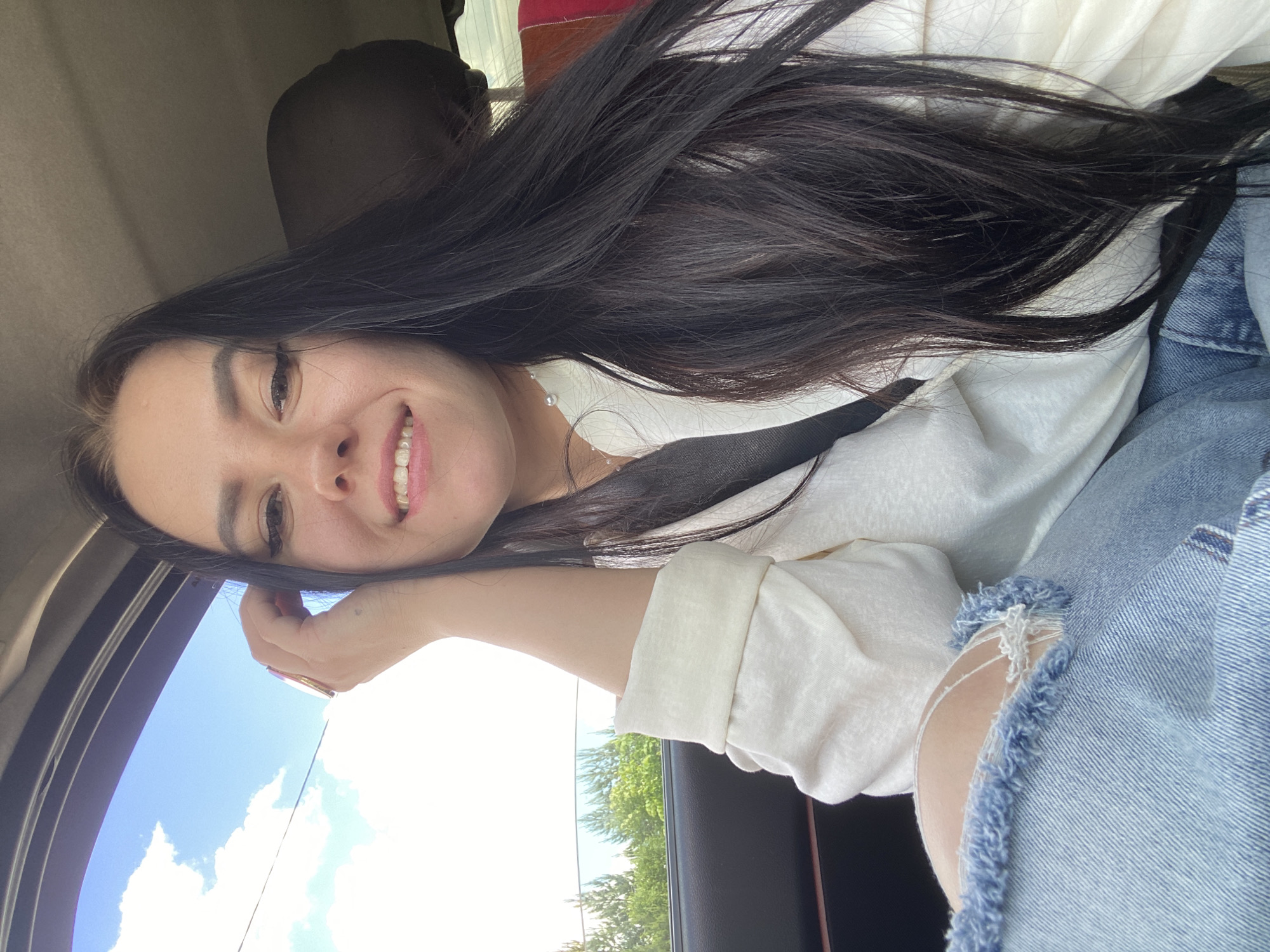 A woman with long, dark hair is sitting in a car, smiling at the camera. She's wearing a white top and light blue jeans with a rip in the knee. A seatbelt is visible across her chest. Outside the car window, there's a view of a bright blue sky with white clouds, and green trees can be seen in the distance. The location is likely Votkinsk, Russia, but there is no information in the picture to confirm that location.
