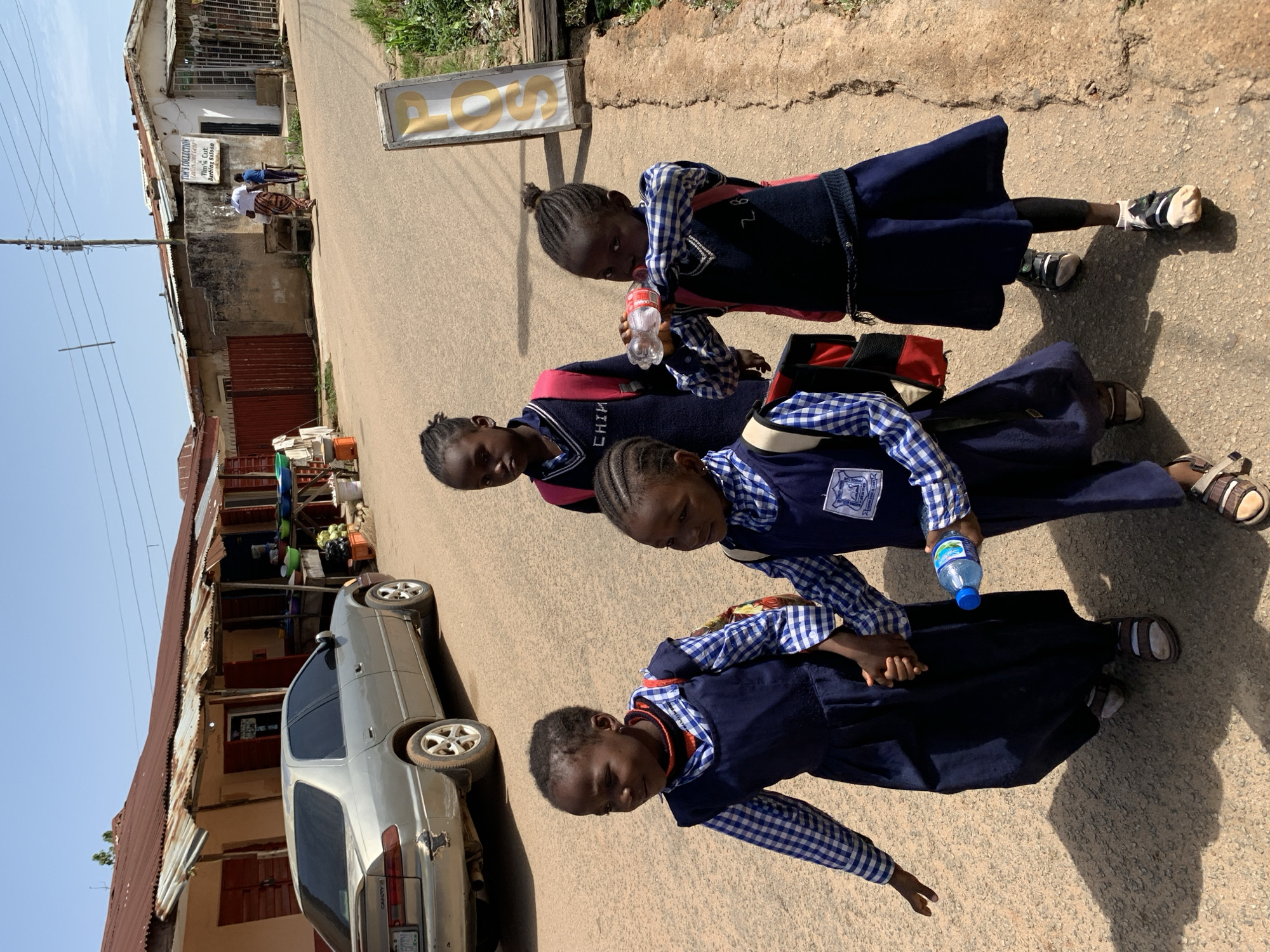 The image depicts a group of four young girls in school uniforms walking on a paved street in the city of Jos, Nigeria. The girls are wearing dark blue jumpers over blue and white checkered shirts, with some also wearing dark sweaters or vests. Three of the girls are in the foreground, with the fourth slightly behind them. The girl in the middle of the foreground is holding the hand of the girl to her left, and the girl to her right is drinking from a bottle of water. The background shows a street lined with buildings that appear to be shops or residences, with corrugated metal roofs. A silver car is parked on the left side of the street. The sky is clear and the sun is shining, casting shadows on the ground. A signpost with the letters "POS" is visible on the right side of the image. The environment suggests a bustling urban or semi-urban area.
