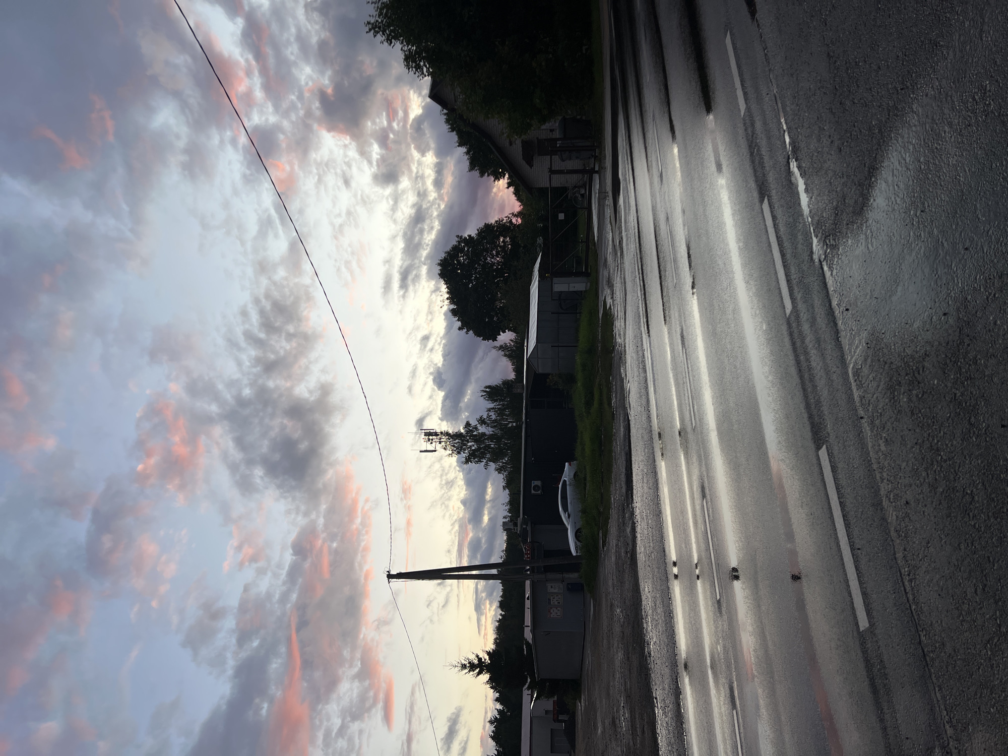 The image depicts a wet asphalt road in Vilnius, Lithuania, during a cloudy sunset. The sky is a mix of blue and grey clouds with streaks of pink, indicating the sun is low on the horizon. A telephone pole stands on the left side of the road, and in the background, there are several industrial-looking buildings, a white car, trees, and a tall communication tower. The road surface is reflective, showing puddles and the white lane markings. The overall atmosphere is dim and moody, characteristic of a twilight scene after rain.