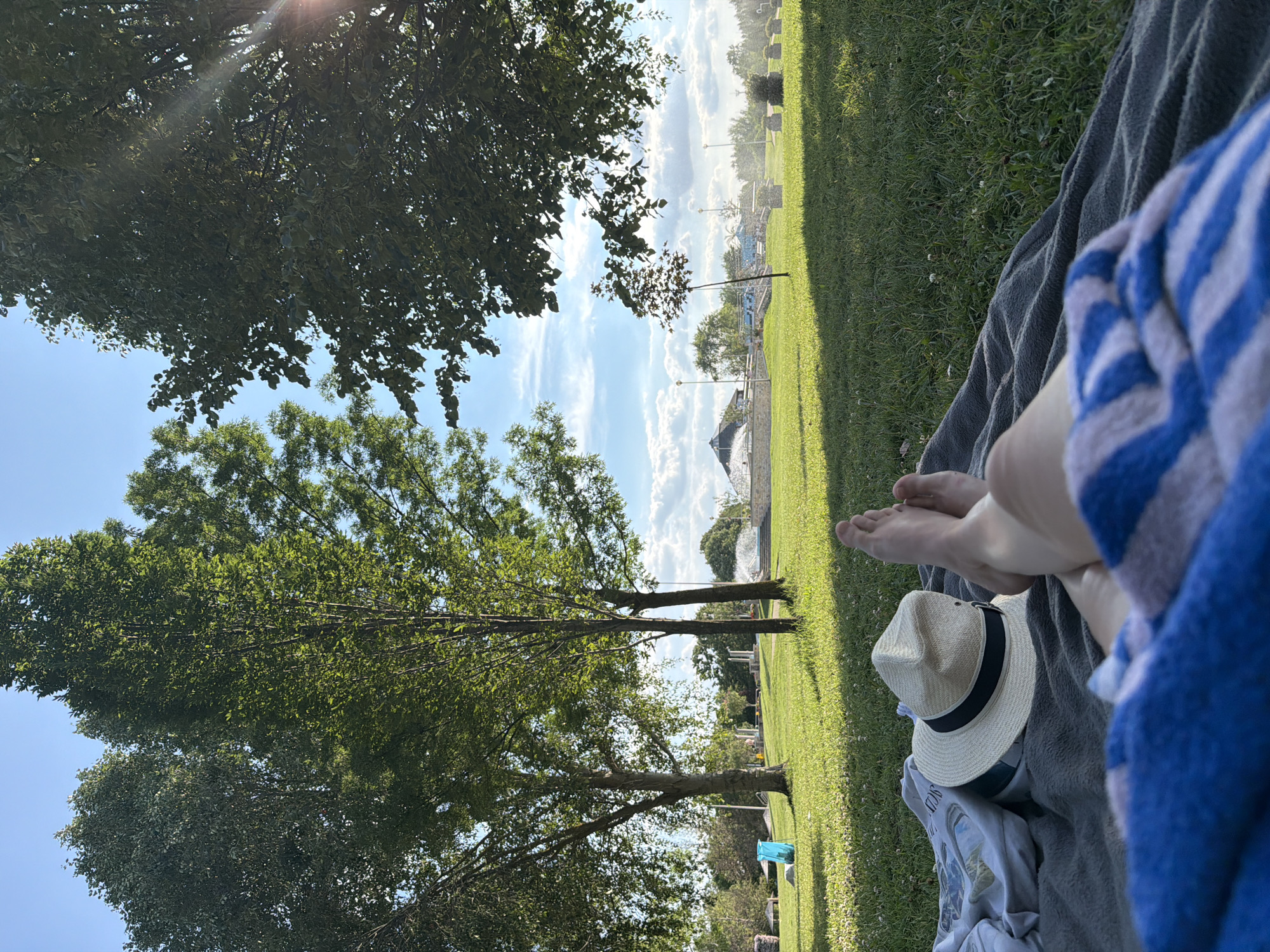 The image depicts a person relaxing on a blanket in a grassy park on a sunny day. In the foreground, the person's legs and feet are visible, covered partially by a blue and white striped towel. A straw hat with a black band rests on a gray blanket next to a crumpled white garment.
The background shows a well-maintained park with lush green grass, several trees with dense foliage, and a bright blue sky with scattered white clouds. In the mid-ground, there are structures that appear to be part of a public facility, possibly a water park or a fountain area, with water spraying in the distance. Lampposts and low-lying stone planters are also scattered throughout the park. A bright blue plastic bag or container is visible on the grass to the left of the frame. The overall impression is one of leisure and outdoor recreation in an urban park setting.
While the image provides a visual representation of a park scene, it does not contain any specific information or landmarks that would allow for definitive identification of the location within the city of Kiskunmajsa, Hungary. The scene is generic to many parks found in temperate climates during warmer months.