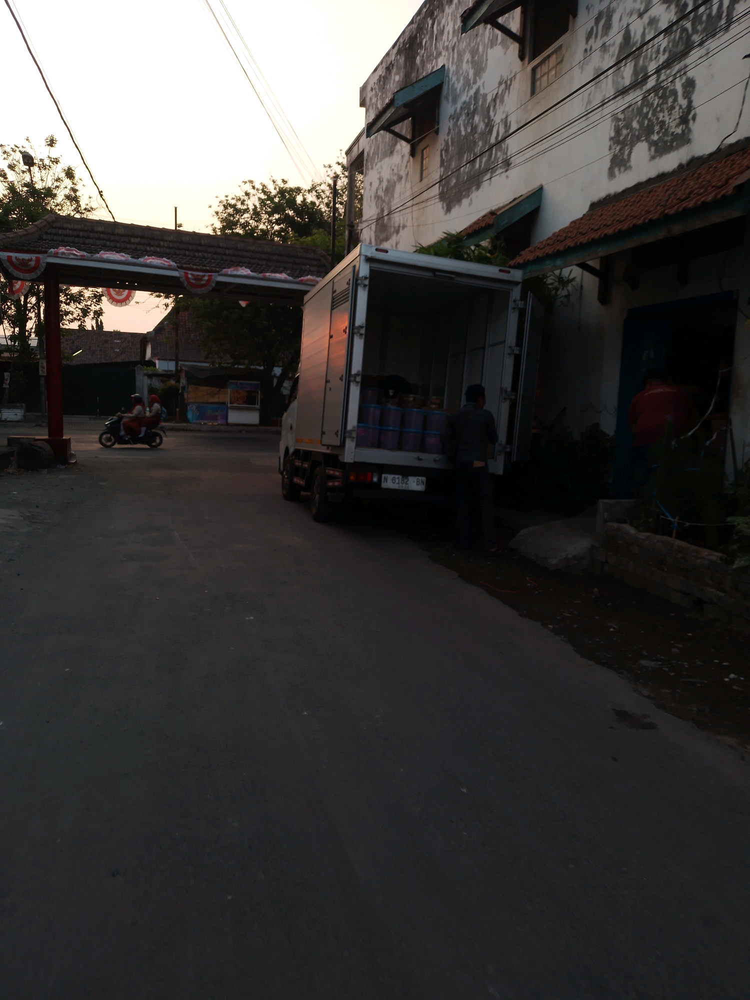 A cargo truck with its rear doors open is parked on a paved road in Kertosono, Indonesia, near a building with peeling paint. Two men are standing by the truck, one with his back to the camera and the other wearing a red shirt. In the background, a motorcycle with two people on it is riding down the road, and a red and white decorative archway is visible, possibly for a celebration. The sky is overcast, suggesting it might be early morning or late afternoon. The scene depicts a moment of activity in a street setting, with elements of commerce and daily life.