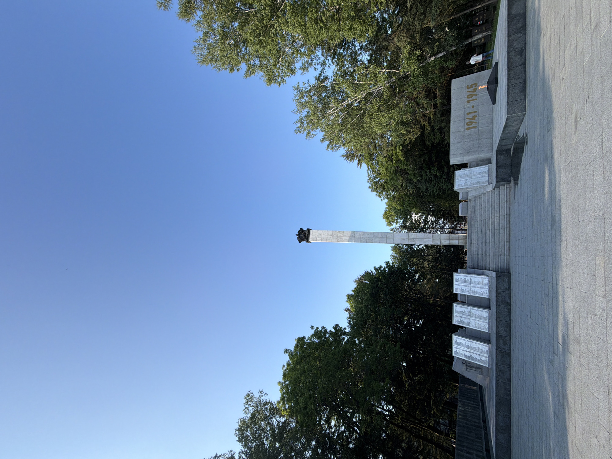 The image depicts the Eternal Flame memorial in Birobidzhan, Russia, located on a plaza. A tall, marble obelisk topped with a dark structure stands prominently in the center, flanked by trees. In front of the obelisk, a set of stairs leads up to the memorial. To the left of the stairs, several white plaques with lists of names are displayed at an angle. To the right of the stairs, a large concrete structure bears the inscription "1941-1945" in gold lettering, with an eternal flame burning in front of it. A single individual, a person wearing a white shirt and blue jeans, stands near the eternal flame, facing away from the camera and towards the trees. The sky is clear and blue. The plaza is paved with large, light-colored square tiles. Lush green trees line the background, partially obscuring other structures and possibly tombstones further back on the right.