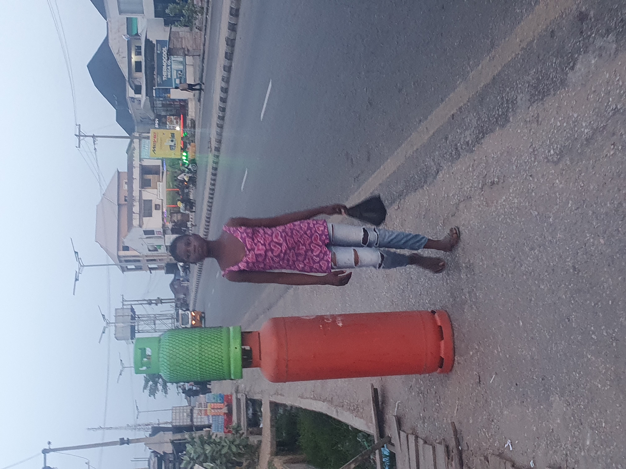 The image shows a person lying prone on the shoulder of a paved road in Lokoja, Nigeria. The person is wearing a pink floral print top and ripped blue jeans, and is holding a black hat. Two gas cylinders, one orange and one green, lie adjacent to the person. The road has a yellow painted line. Buildings and utility poles are visible in the background.