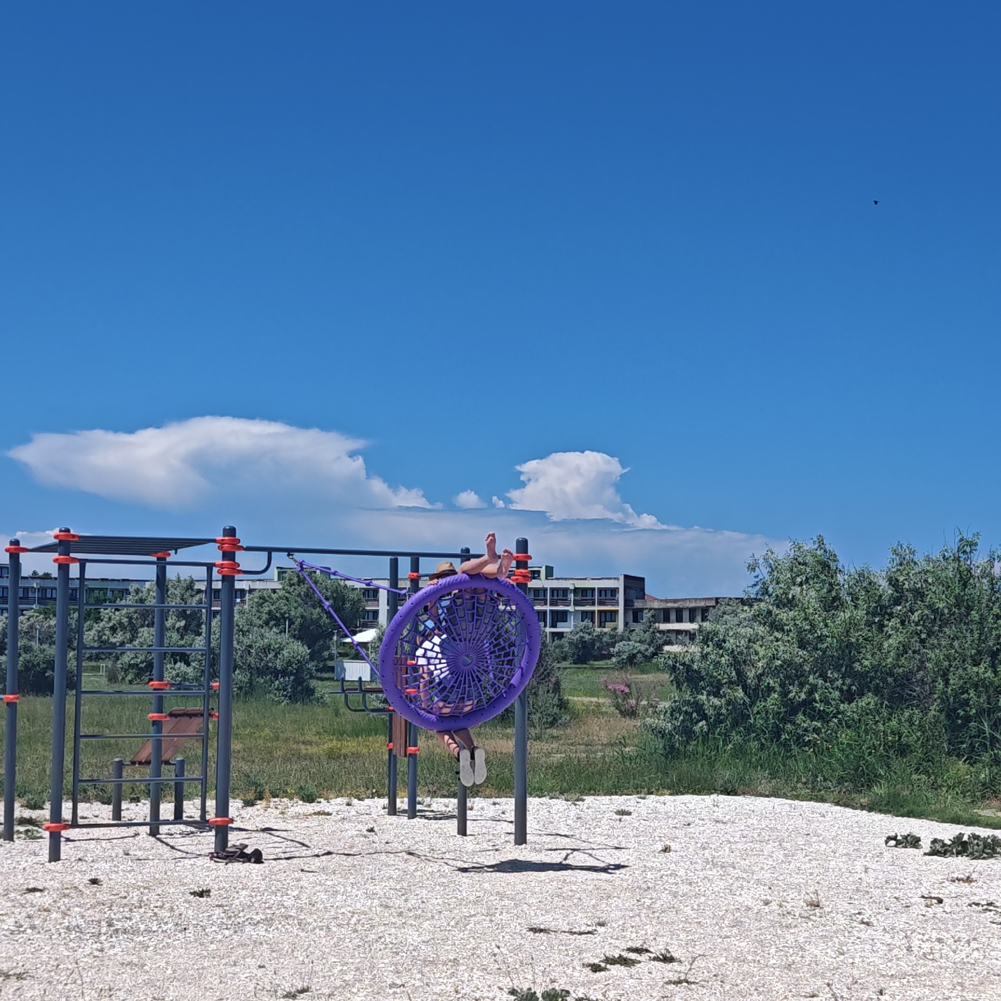 This image depicts a scene on a playground, likely in an outdoor recreational area, with a bright blue sky and scattered clouds. A person, only their lower body visible, is seated on a large purple nest-like swing. Their feet, clad in light-colored shoes, are extended outwards, and their legs are bent at the knees. The swing is suspended from a metal frame which is part of a larger playground structure, also featuring horizontal bars and what appears to be a climbing ladder. The ground beneath the playground equipment is covered in small white pebbles, with patches of green grass and some low-lying vegetation interspersed. In the background, there are buildings, some appearing to be hotels or apartment complexes, situated behind a dense line of green trees and shrubs. The overall impression is of a sunny day at a seaside resort or a similar coastal location, given the flat terrain and the type of vegetation.