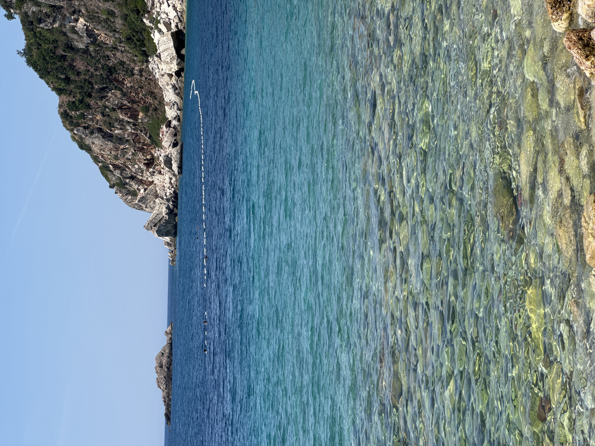 The image depicts a serene coastal scene at Perazića Do, Montenegro, with the camera oriented vertically, making the water appear to be flowing from left to right across the frame. The left side of the image features a rugged, rocky cliff face covered in green vegetation, sloping down towards the sea. A clear blue sky is visible above the cliff.
The water itself is the dominant subject, transitioning from a deep blue hue near the shore to a more transparent, turquoise color further out. The transparency of the water allows the rocky seabed and scattered stones beneath the surface to be clearly visible, creating a mesmerizing, rippled effect.
Along the boundary between the darker and lighter blue water, a white, dashed line of buoys stretches across the frame, likely marking a swimming area or a boundary for water activities. Several dark, small objects, possibly swimmers or small boats, are visible in the water near the buoys.
The overall impression is one of a calm and clear day, likely mid-morning or early afternoon given the bright sunlight reflecting off the water and illuminating the seabed. The location is identifiable by the distinctive rocky cliffs and the characteristic clear, blue waters of the Montenegrin coast. There is no visible text in the image.