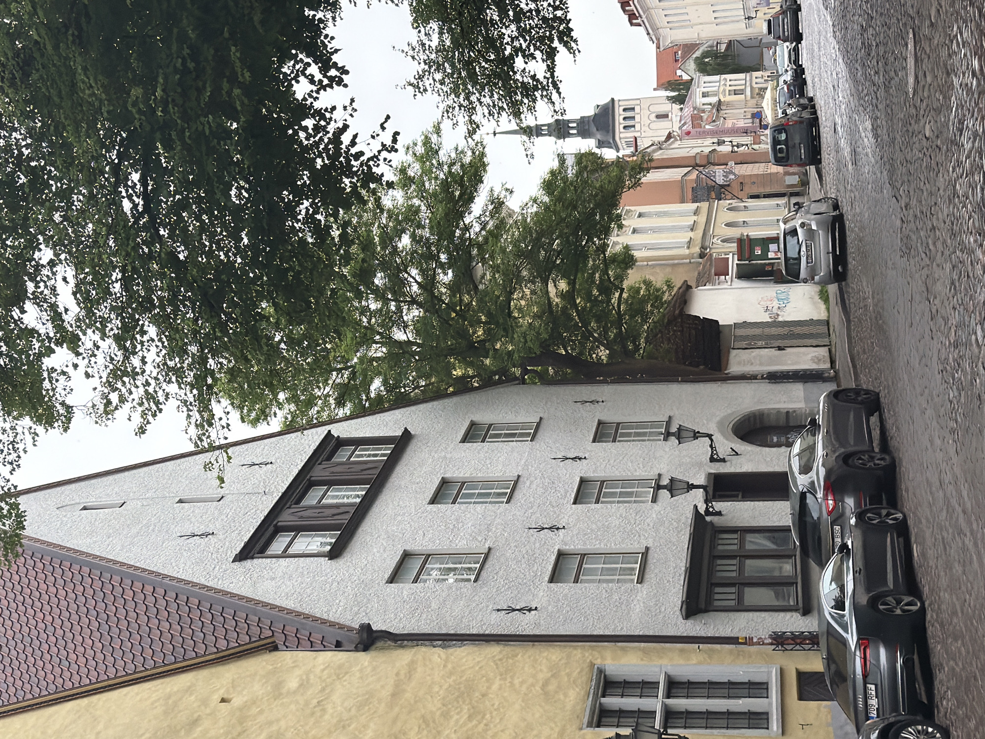 The image shows a street scene in Tallinn, Estonia. The foreground features a cobblestone street lined with parked cars, including a silver Audi and a dark gray Maserati. On the left, a tall, narrow building with a yellow facade and a steeply pitched tiled roof is visible. Adjacent to it is a white building with a similar steep roof, featuring several windows of varying sizes and decorative ironwork on its facade.
In the middle ground, more buildings with colorful facades line the street, interspersed with trees that cast shadows. A prominent church tower with a distinctive spire rises above the rooftops in the distance. The street appears to be a pedestrian zone or a street with limited vehicle access, given the presence of parked cars and the cobblestone pavement. The sky is overcast, suggesting a cloudy day. The overall atmosphere is that of a historic European city.