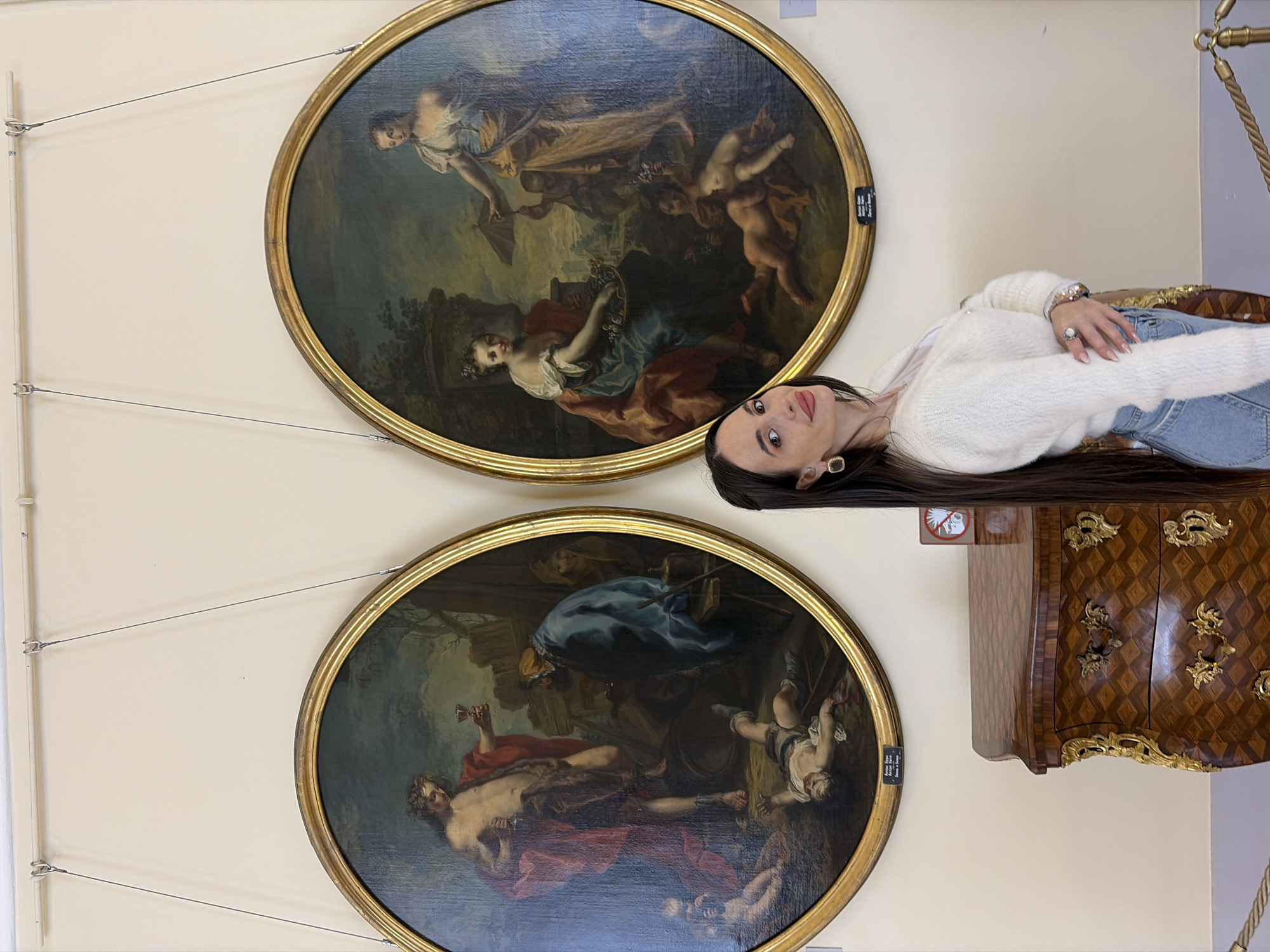 A woman is standing in a museum, with two round paintings on the wall behind her. She is wearing a white sweater and jeans, with long dark hair and jewelry. The paintings appear to be old master style, depicting classical scenes with figures and landscapes. In the lower painting, a man is reclining with a glass of wine and another figure is falling from a chariot. The museum is likely located in Saint Petersburg, Russia. A wooden cabinet is also visible to the woman's right.