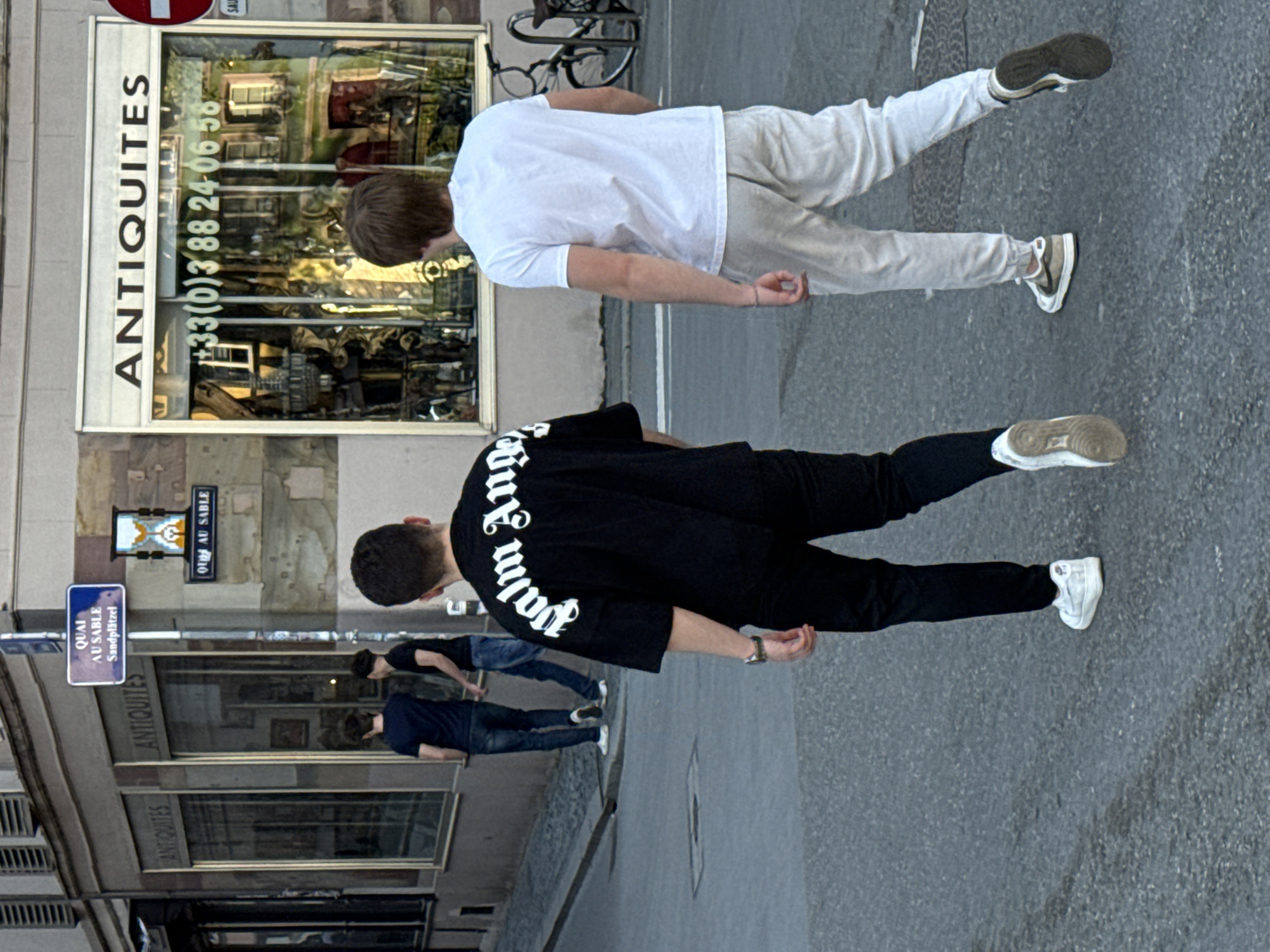 An urban street scene, likely in Strasbourg, France, is depicted during daylight hours with a clear sky. Two young men are walking away from the viewer on a paved road. The man in the immediate foreground wears a black t-shirt featuring white text, partially reading "palm Ange," along with black trousers and white sneakers. Next to him, another man wears a plain white t-shirt, light beige or white trousers, and light-colored sneakers with dark soles. Both appear to be casually strolling.
On the left side of the frame, buildings with large shop windows reflect their surroundings. A prominent shop is identified by the vertical text "ANTIQUITES" on its frame and a phone number "+33(0)3 88 24-06 58" on the glass, indicating an antiques store. Further down the street, additional text reading "ANTIQUITES" is visible on another shop. Street signs clearly display "QUAI AU SABLE" and "Sandplätzel," which is consistent with the bilingual nature of Strasbourg. A bicycle rack is partially visible between the two main subjects and the shop window. In the background, two other individuals are seen near a building entrance. The overall scene suggests a typical daytime street environment, with no discernible emotions from the subjects.