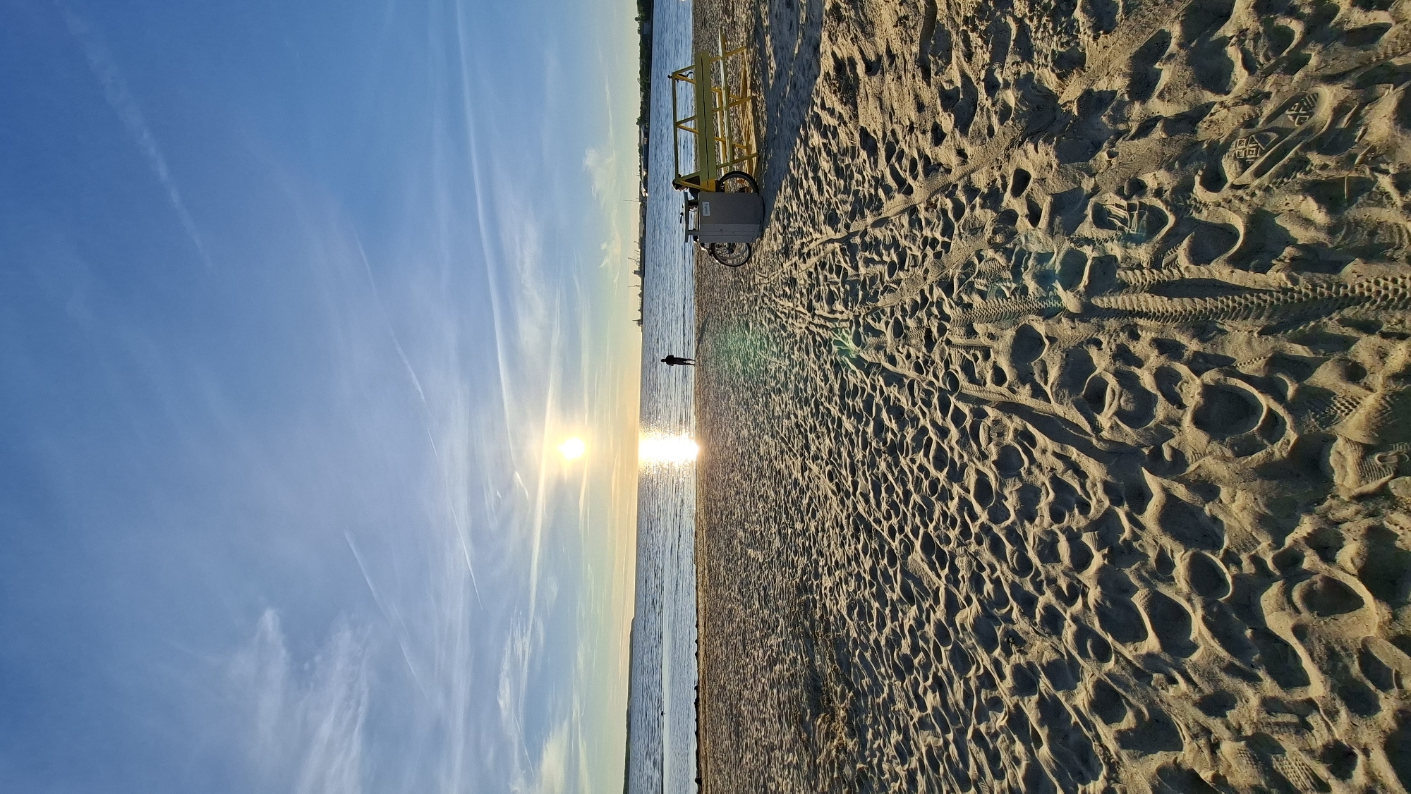 The image depicts a beach in Tallinn, Estonia, at sunset. The sun is low on the horizon, casting a warm glow across the water and the sandy shore. The sea is calm, with a few distant boats visible on the horizon. The beach is mostly empty, with a single person standing near the water's edge. There is a yellow bench and a bicycle parked on the sand to the right of the frame. The sand is imprinted with tire tracks and footprints, suggesting recent activity. The sky is a clear blue, with a few wispy clouds.