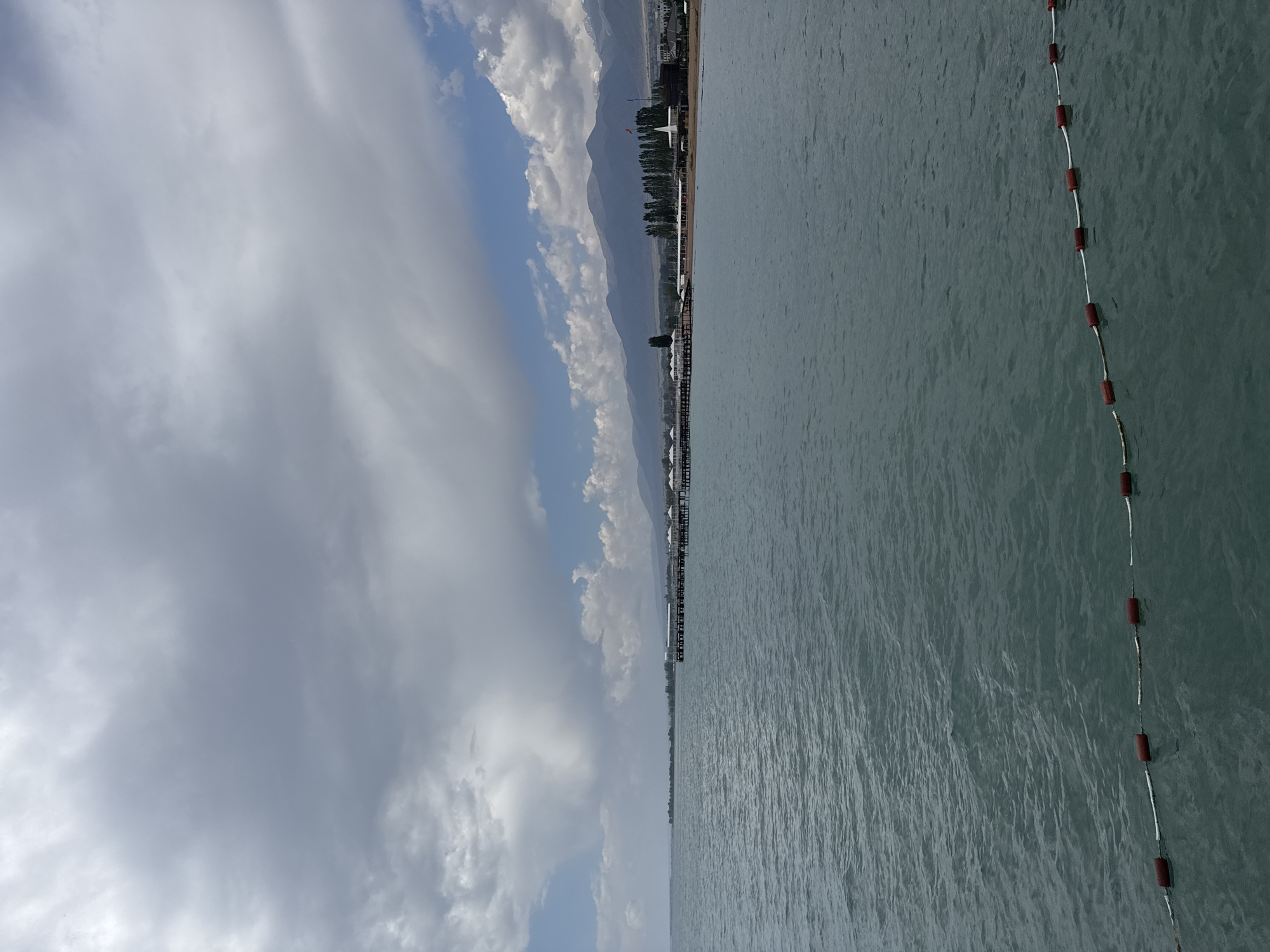 A wide-angle, low-angle shot captures a serene lake scene in Sary-Oy, Kyrgyzstan, under a dynamic, cloudy sky. The foreground is dominated by the calm, blue-green water of the lake, with a marker buoy line of red floats and white rope stretching across the lower part of the frame. In the mid-ground, a long, wooden pier extends into the lake, lined with small, white, tent-like structures. Beyond the pier, a tree-lined shore is visible, with a section of beach and buildings. In the background, a range of majestic, hazy mountains rises, their peaks partially obscured by the clouds. The overall atmosphere is peaceful and expansive.