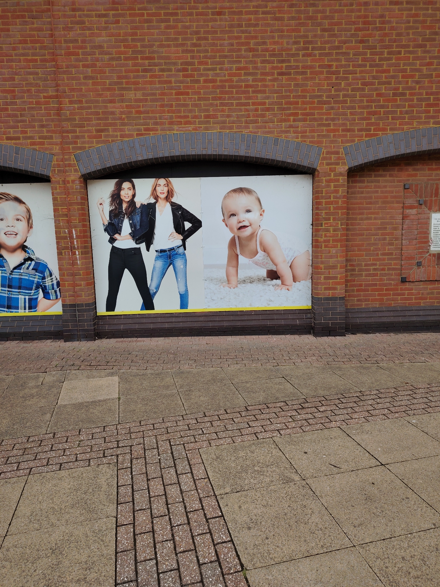 The image depicts the lower section of a brick building facade fronting a paved sidewalk, likely located in Peterborough, United Kingdom.
The building is constructed from red bricks with yellowish mortar, featuring three ground-level display windows. The window frames are obscured by the depicted images, but dark grey-blue brick arches are visible above each window recess. Between the left and middle window, and the middle and right window, are vertical red brick columns with dark grey-blue brick bases. On the far right, a segment of the red brick wall contains a recessed square panel of bricks secured by four black metal brackets. Below this panel, a faint white plaque is visible with partially legible text, including "THE CAMBRIDGESHIRE REGIMENT" and a list of names such as "RICHARD ANTHONY", "HERBERT EDWARDS", and "JAMES KILBY". This historical plaque, referencing the Cambridgeshire Regiment, indicates a strong connection to Cambridgeshire and supports the location being in Peterborough.
The display windows feature large photographic advertisements:
* The leftmost visible panel shows the upper body of a smiling young boy with light hair, wearing a blue and white plaid shirt.
* The central panel displays two adult women standing side-by-side. The woman on the left has dark hair and wears a denim jacket over a white top and black pants. The woman on the right has lighter hair and wears a black leather jacket over a white top and blue ripped jeans.
* The rightmost visible panel depicts a smiling baby with light hair, crawling on a white fluffy rug while wearing a white onesie.
The sidewalk in the foreground is paved with large, light brown/grey rectangular concrete slabs. A patterned strip of smaller, reddish-brown speckled pavers runs horizontally across the mid-ground, with a perpendicular section extending towards the bottom right of the frame, indicating a pathway or decorative element.