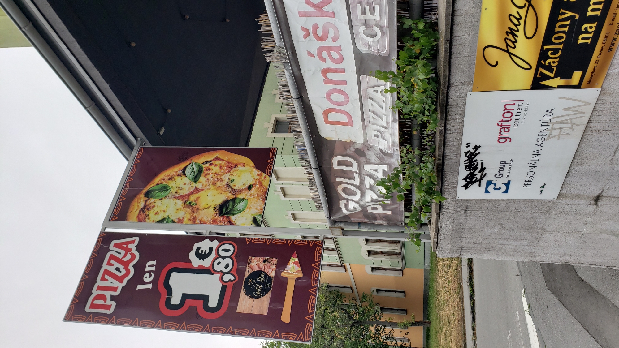The image captures a street scene in Prešov, Slovakia, featuring a series of signs mounted on a building's exterior and a concrete wall. Dominating the upper left is a maroon banner advertising "PIZZA" with the price "1 €80" clearly displayed, alongside an image of a pizza. Adjacent to it, another banner showcases a close-up of a pizza with basil. Below these, a banner reads "GOLD PIZZA". Further to the right, a banner displays the word "Donášková" in large pink letters, followed by "PIZZA" and "CE". On a concrete wall in the foreground, a white sign is visible with logos for "Group" and "grafton recruitment", and text indicating "PERSONALNA AGENTURA". Another yellow sign to the right advertises "Jana" and "Záclony a na m". The background reveals buildings with pastel-colored facades and visible windows. Vegetation, including leafy branches, grows along the top of a fence or railing in front of the signs on the wall. The sky is overcast.