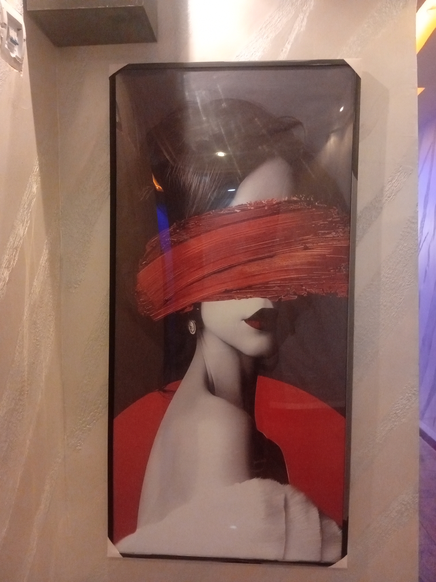 A framed artwork is displayed on a wall. The artwork is a portrait of a woman with her eyes covered by a broad stroke of red paint. She is depicted from the shoulders up, with her head tilted slightly to the left. The background of the artwork is dark, with some subtle lighting effects. The wall behind the artwork has a textured finish in a neutral color. Above and to the left of the artwork, a portion of a shelf or ledge is visible. The overall impression is that of a modern art piece in a domestic or commercial setting. The location is identified as Sabon Gari East, Nigeria.