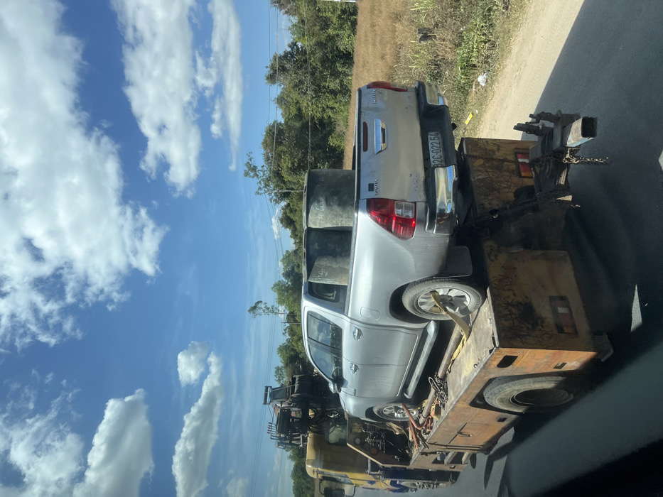 A silver Toyota Hilux sits on the back of a flatbed tow truck. The pickup truck contains cylindrical objects in its bed. The tow truck, primarily yellow, shows signs of rust and wear. A license plate is visible on the rear of the Hilux. The sky is blue with scattered clouds. Trees line the background, and a road is on the right side of the frame.