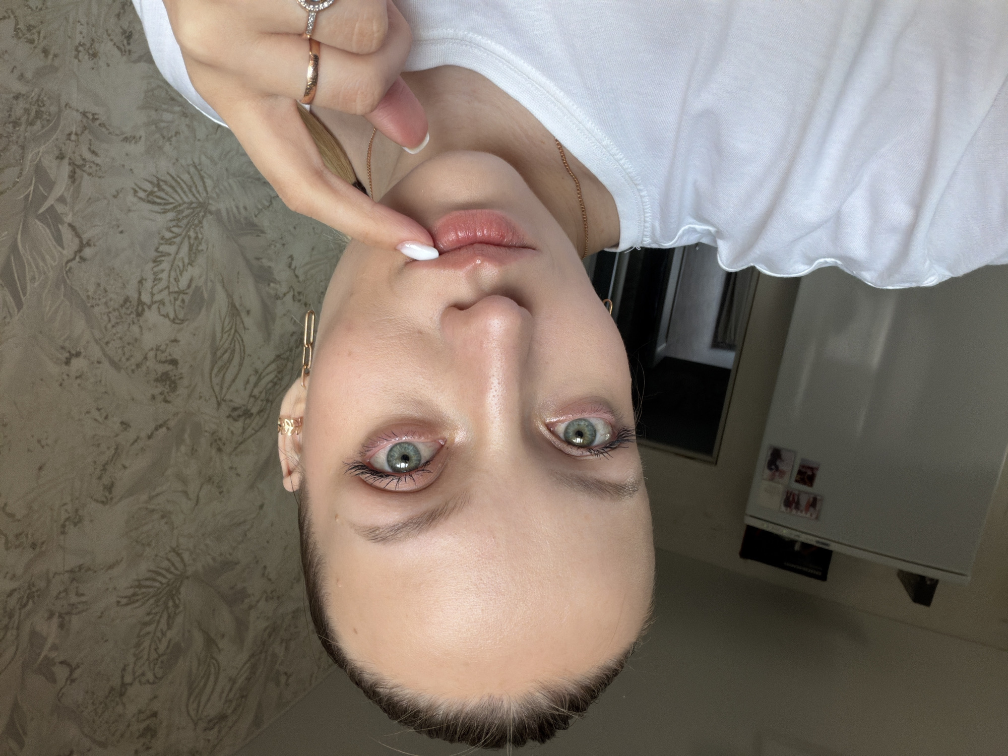 A close-up, front-facing photographic portrait depicts a fair-skinned individual, appearing female, in an indoor residence in Reshetnikovo, Russia. The subject has light green eyes, visible mascara, and light makeup. Hair is pulled back, revealing a high forehead. The subject wears a plain white crew-neck t-shirt, a thin gold-colored necklace, and gold-toned jewelry including multiple rings on the right hand and two distinct earrings in the visible left ear (a small stud and a longer chain-link style). The right index finger, bearing a white-tipped manicure, is positioned against the center of the lower lip. The background on the left shows a portion of a white refrigerator with several small rectangular items attached to its door, and a dark object resting on top. The wall to the right features textured wallpaper with a faint, faded botanical pattern in muted tones. Lighting is ambient, originating from the left side of the frame.