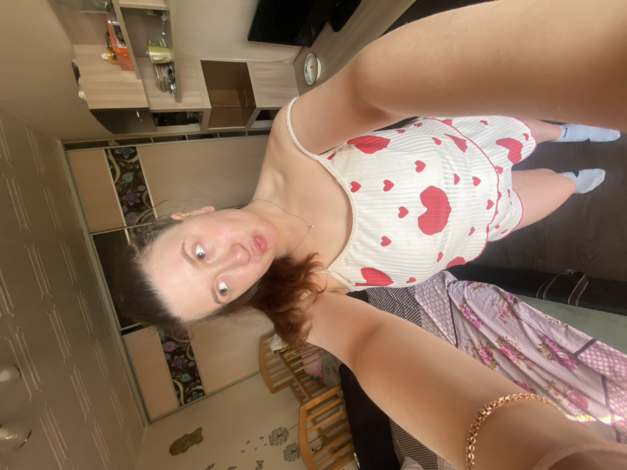 A woman is taking a selfie indoors. She is wearing a white tank top with red hearts and blue socks. A crib is visible in the background. The location may be in Chaikovsky, Russia. The environment appears to be a home. The details suggest a casual setting.