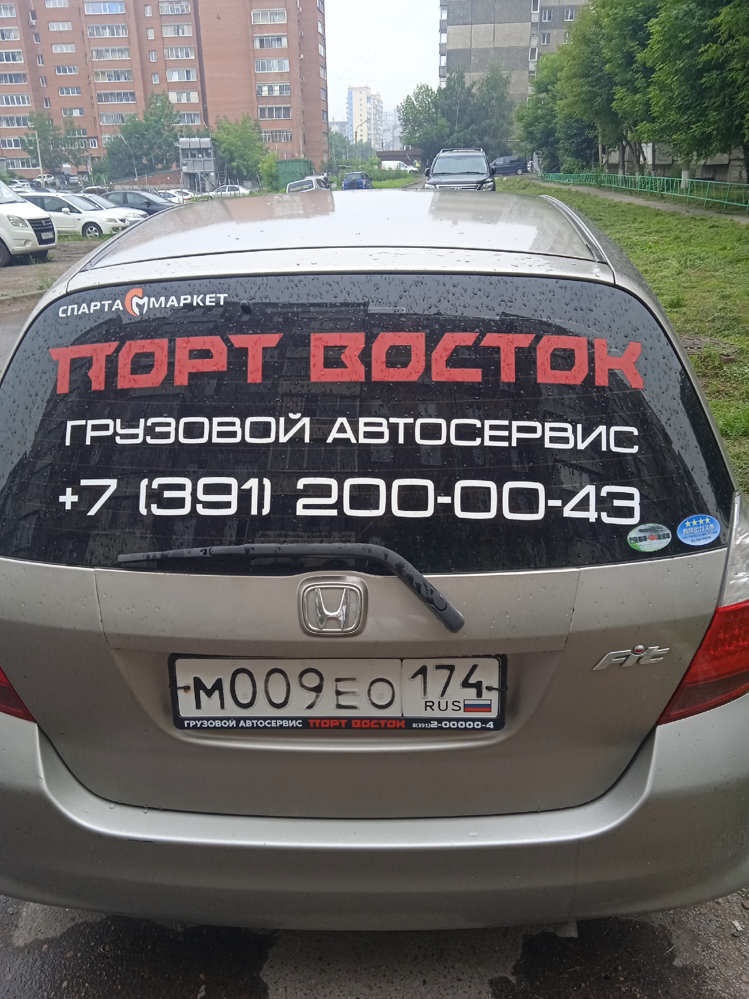 A silver Honda Fit is parked on a wet asphalt surface, possibly in a residential courtyard in Krasnoyarsk, Russia. The car has a clean, slightly worn appearance. Large, red lettering on the rear window reads "ПОРТ ВОСТОК" (Port Vostok), which translates to "Port East." Below this, in white letters, it says "ГРУЗОВОЙ АВТОСЕРВИС" (Cargo Autoservice), followed by a phone number: "+7 (391) 200-00-43". A Russian license plate, "M009EO 174 RUS", is affixed to the rear. The background shows a multi-story brick apartment building, trees, and a green metal fence, suggesting an urban residential setting. The sky is overcast, and the presence of raindrops on the car and ground indicates recent or ongoing precipitation. No people are visible in the frame.